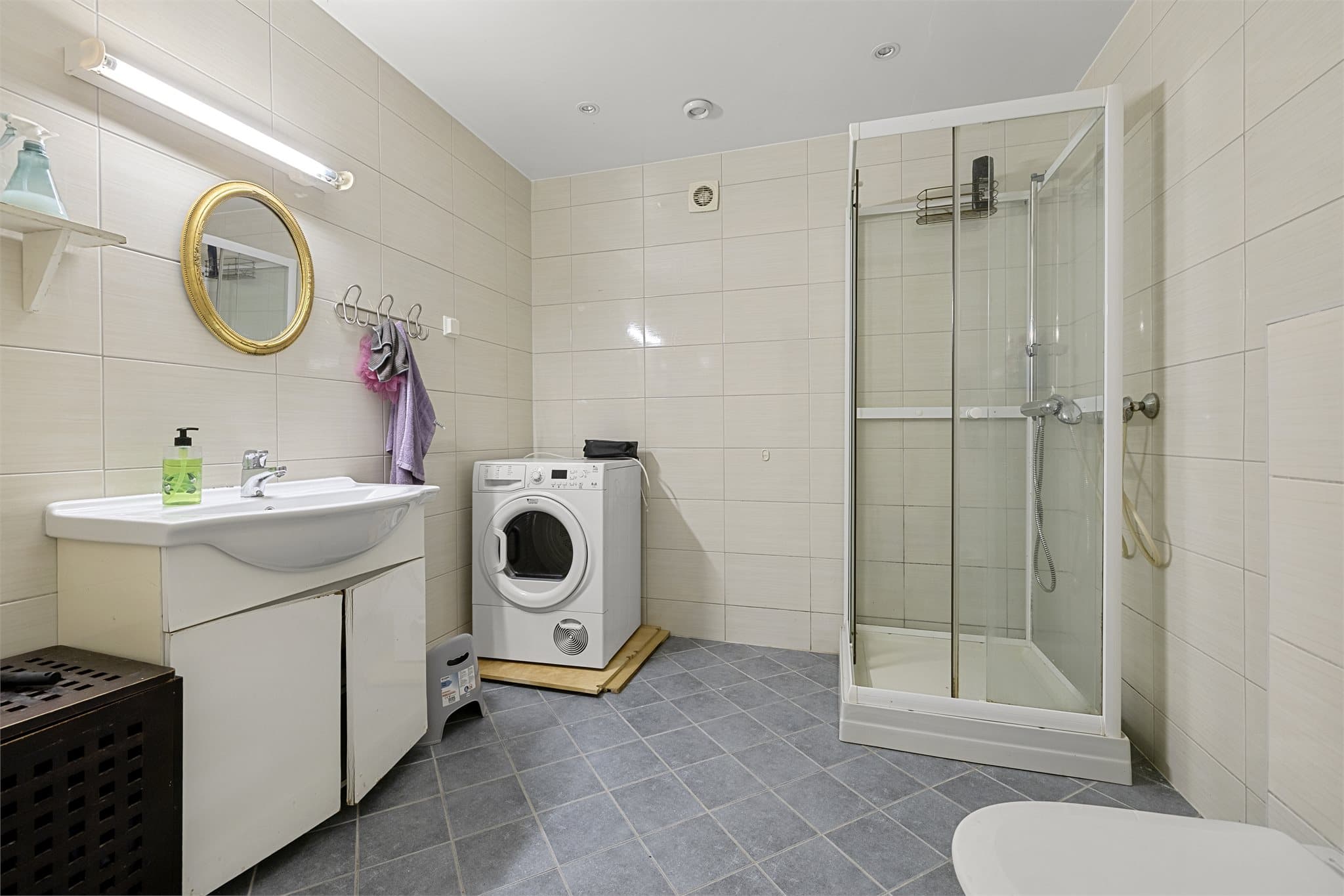 Flislagt bad i kjeller med dusjhjørne, servant og opplegg for vaskemaskin.