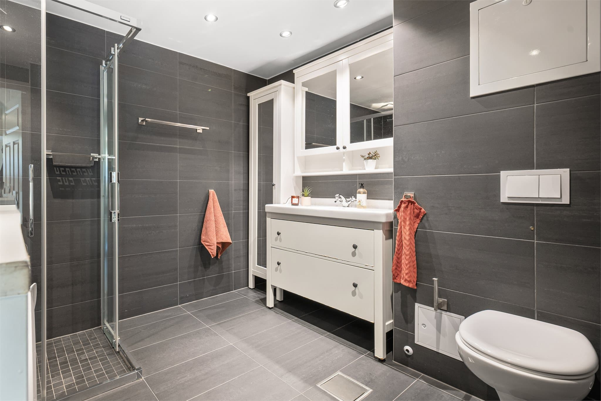 Pent flislagt baderom med tidløse og moderne flisvalg, vegghengt wc, helservant på baderomsinnredning og dusjhjørne med skyvedører i herdet glass.