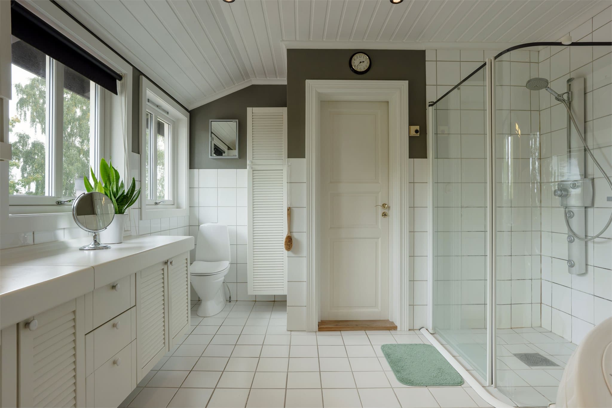 Dusjhjørne med buede glassdører, flislagte overflater og downlights i panelt himling.