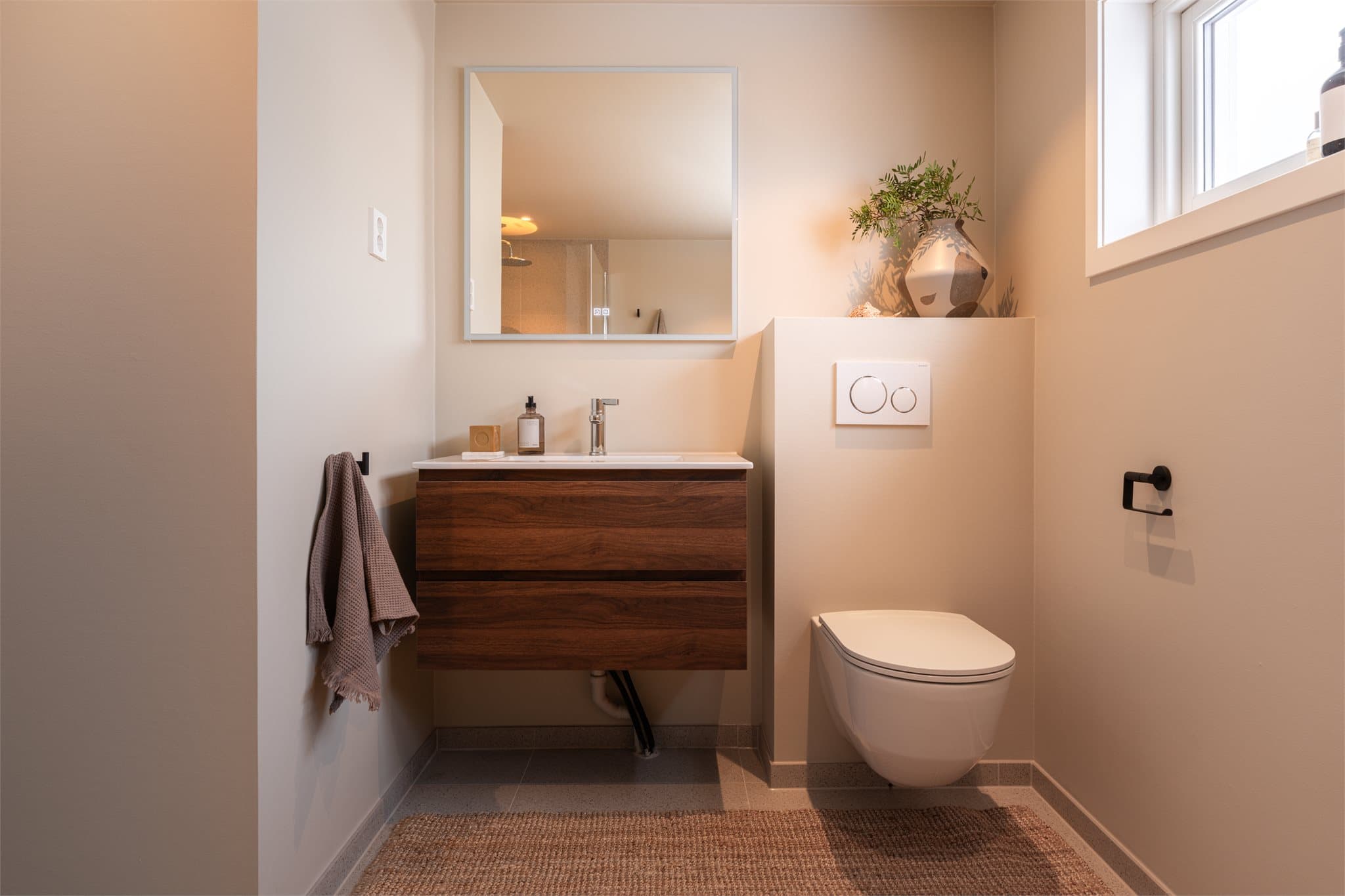 Innredningen består av veggmontert toalett, servant på nøttebrun skuffeseksjon og speil.