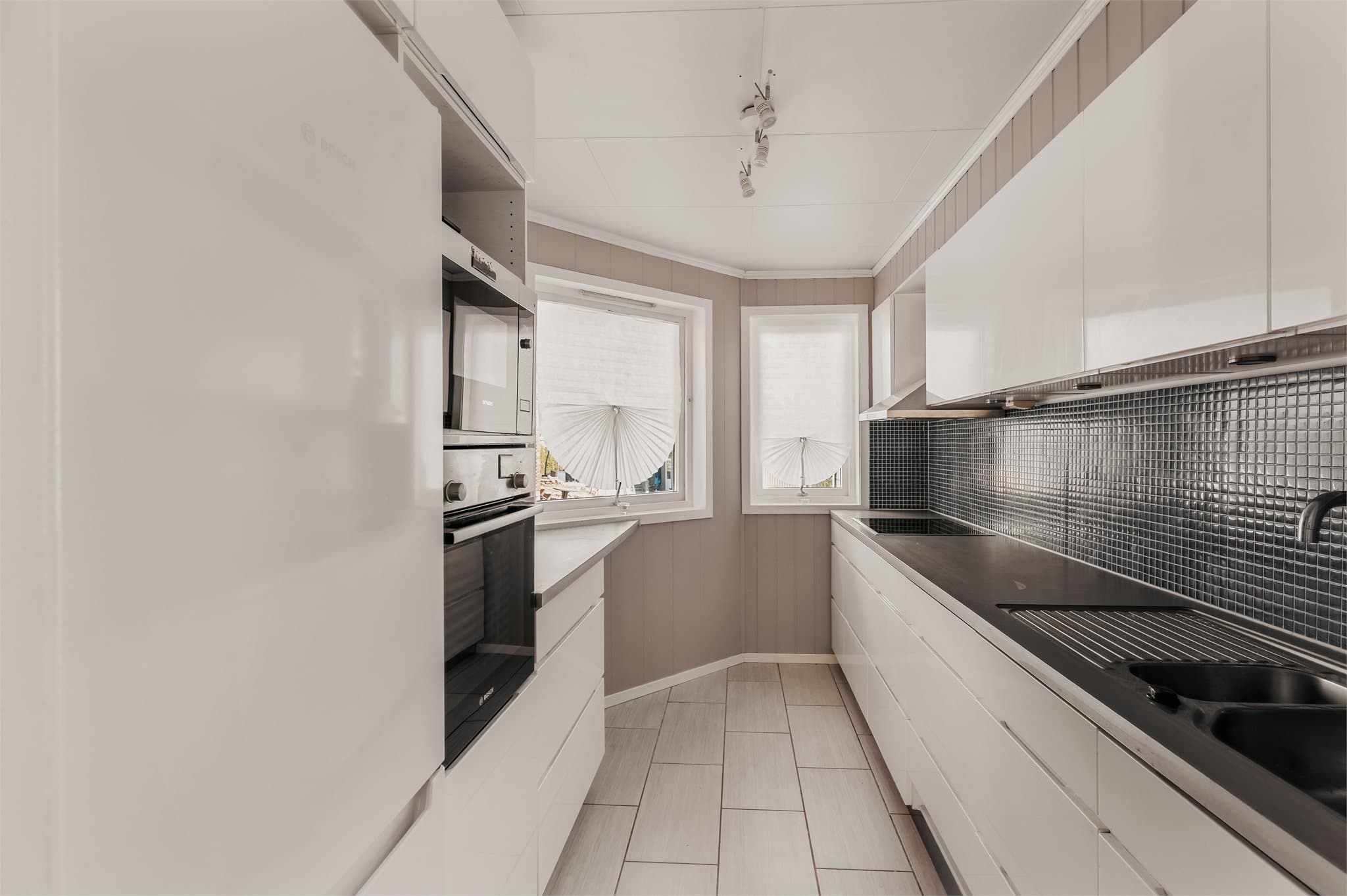 Boligens kjøkkeninnredning har hvite, slette fronter og laminat benkeplate.