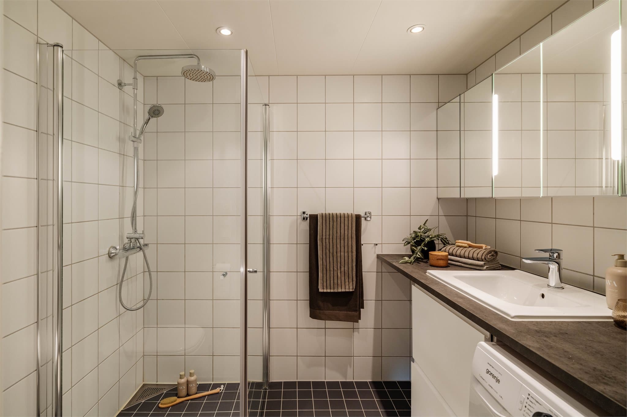 Badet har flislagte vegger og flislagt gulv med vannbåren gulvvarme. Badet består av servant med skuffeinnredning, vegghengt klosett med innebygget sisterne og dusj på gulv med dusjdødrer i herdet glass