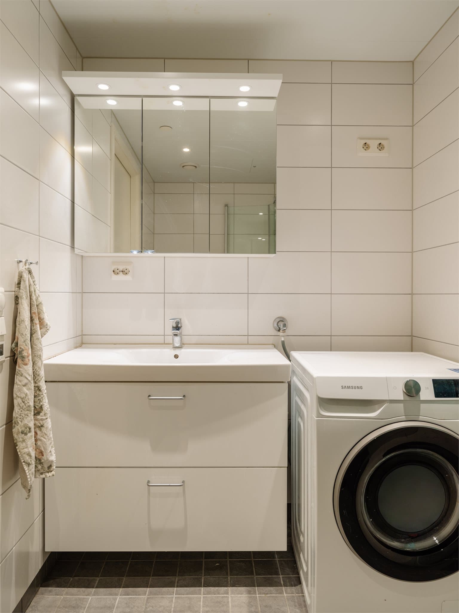 Badet er innredet med dusj med herdet glassdører, servant med skuffeinnredning, vegghengt toalett med innebygget sisterne og opplegg for vaskemaskin