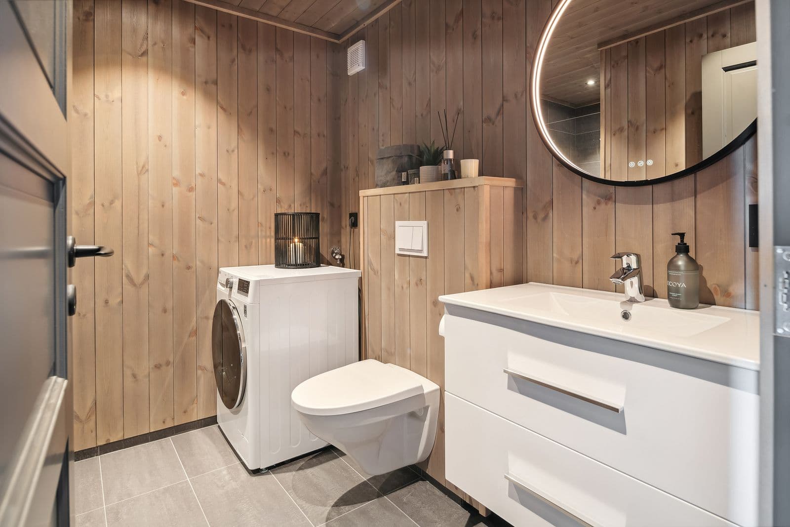 Baderom med delvis panelkledde vegger og gulvfliser med gulvvarme. Vegghengt wc, helservant på skuffeinnredning, speil m/belysning og downlights i himling. Det er opplegg for vaskemaskin.