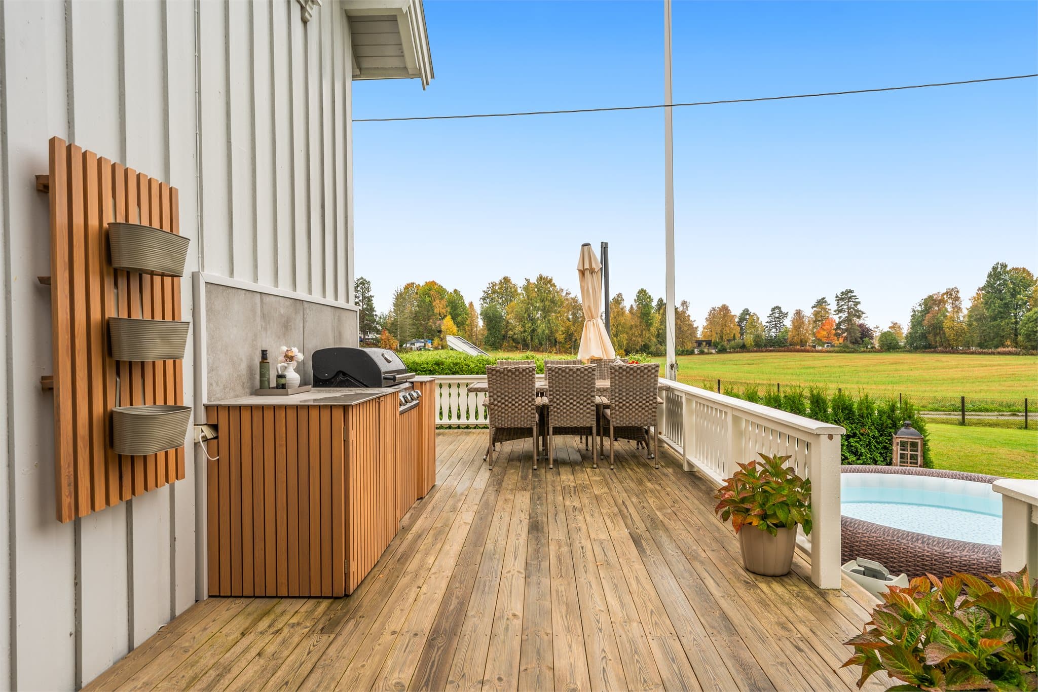 Terrasse i trekonstruksjon med utekjøkken, spiseplass og utsyn mot åkerlandskapet.