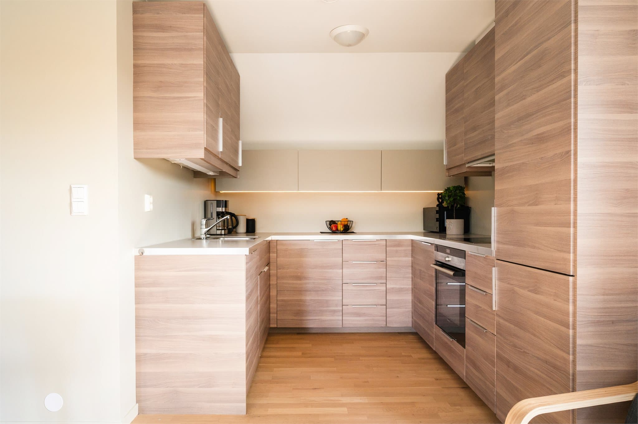 Innredningen fra IKEA har glatte, folierte fronter og benkeplate i laminat med nedfelt kum i rustfritt stål. Det er malte flater på vegg over benkeplate og belysning via lysstoffrør under overskap.