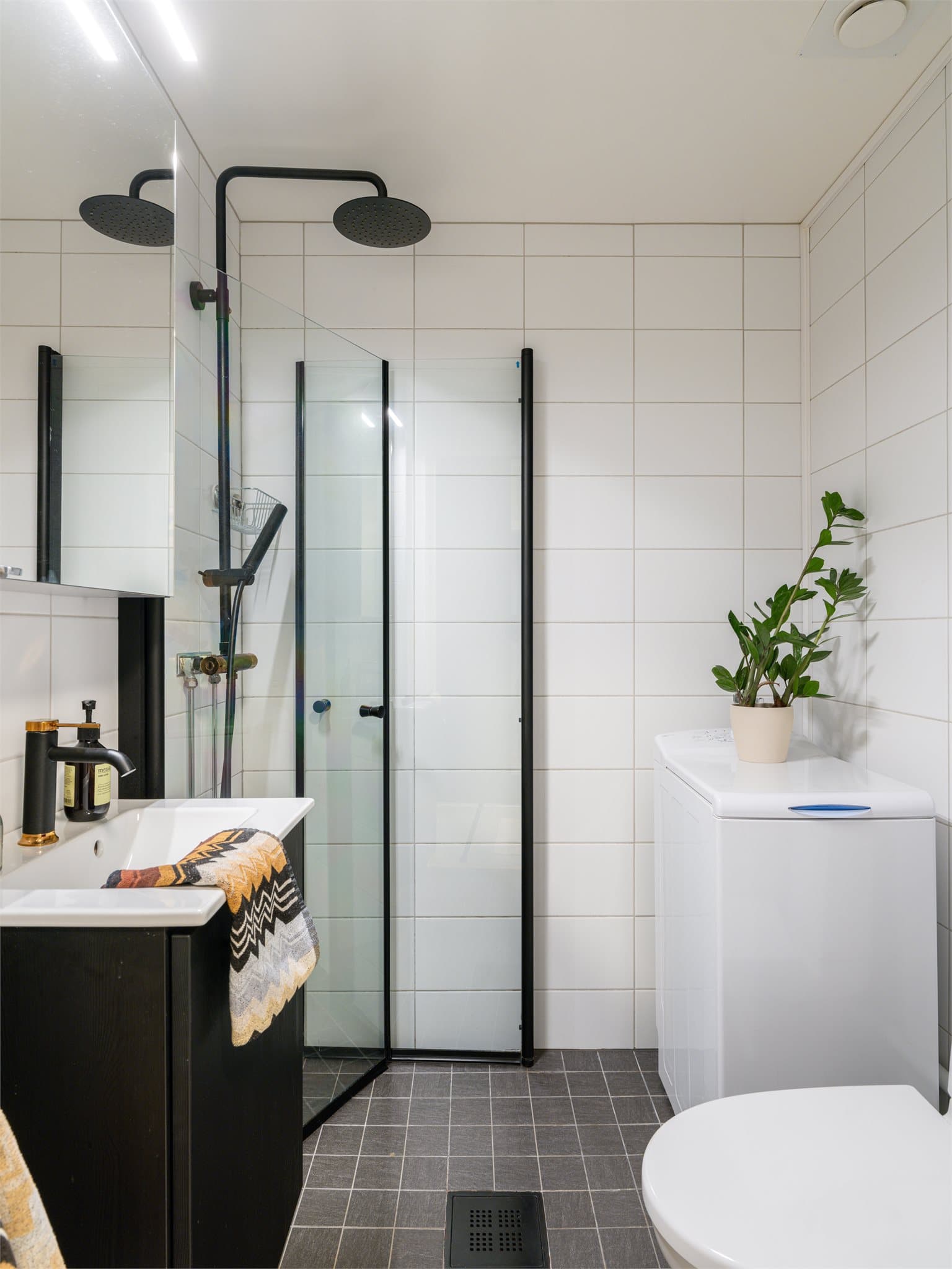 Et lyst og moderne bad med en stilren kombinasjon av hvite veggfliser, grå gulvfliser og sorte detaljer på innredning og armaturer som gir rommet et elegant uttrykk.