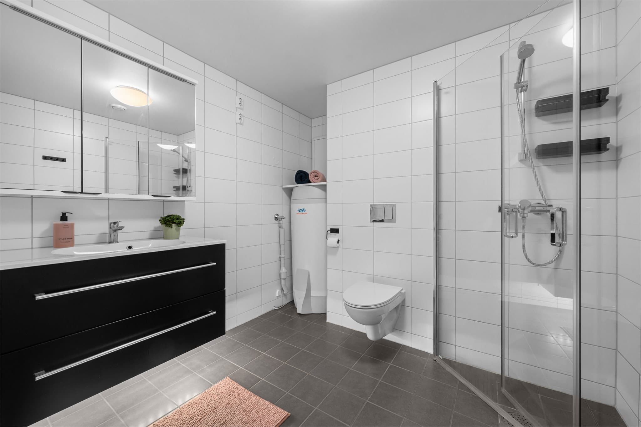 Pent, flislagt baderom med dusjløsning, vegghengt toalett og servant på skuffeseksjon.