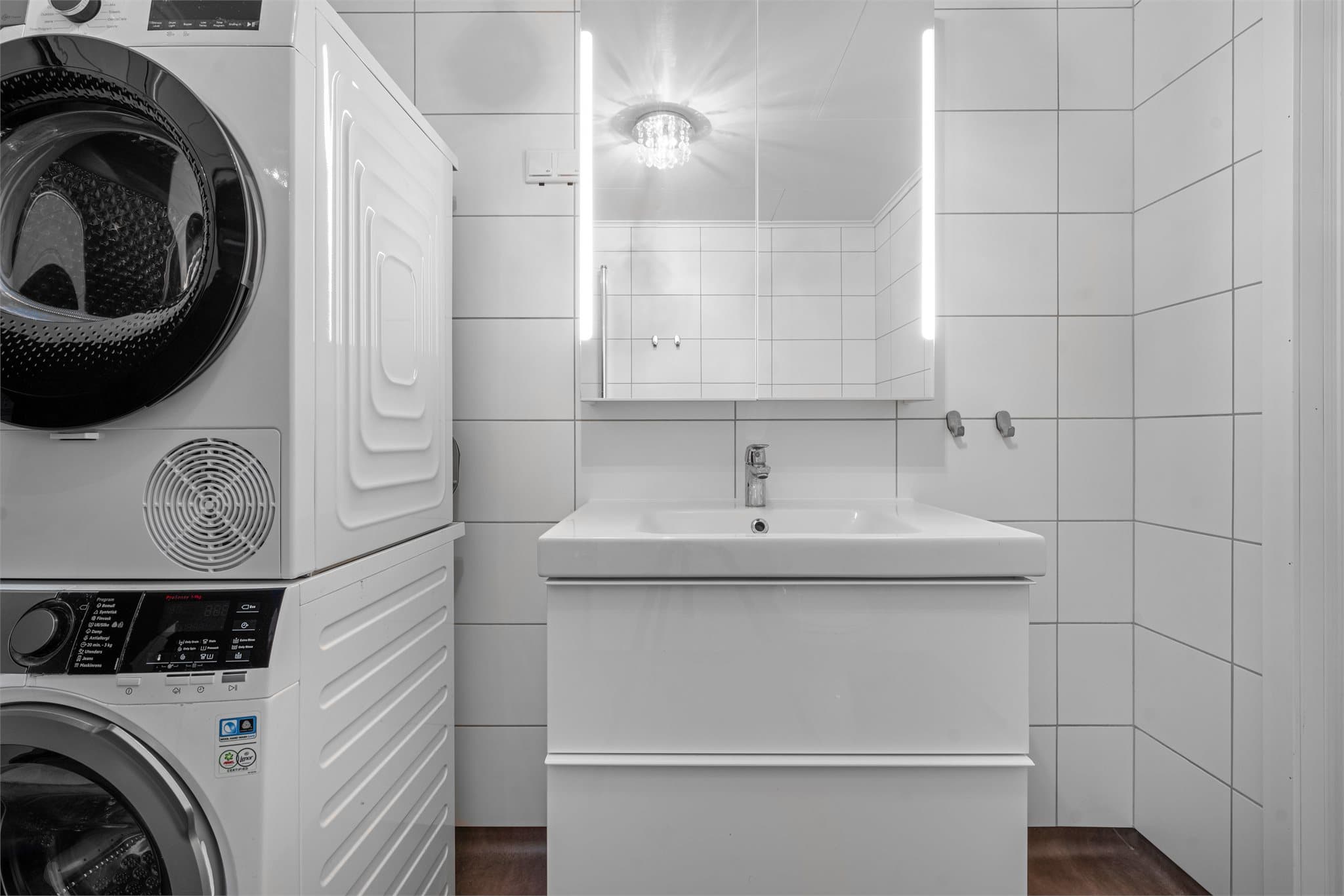 Badet har servant på skuffeseksjon, dusjhjørne, klosett og opplegg for vaskemaskin.