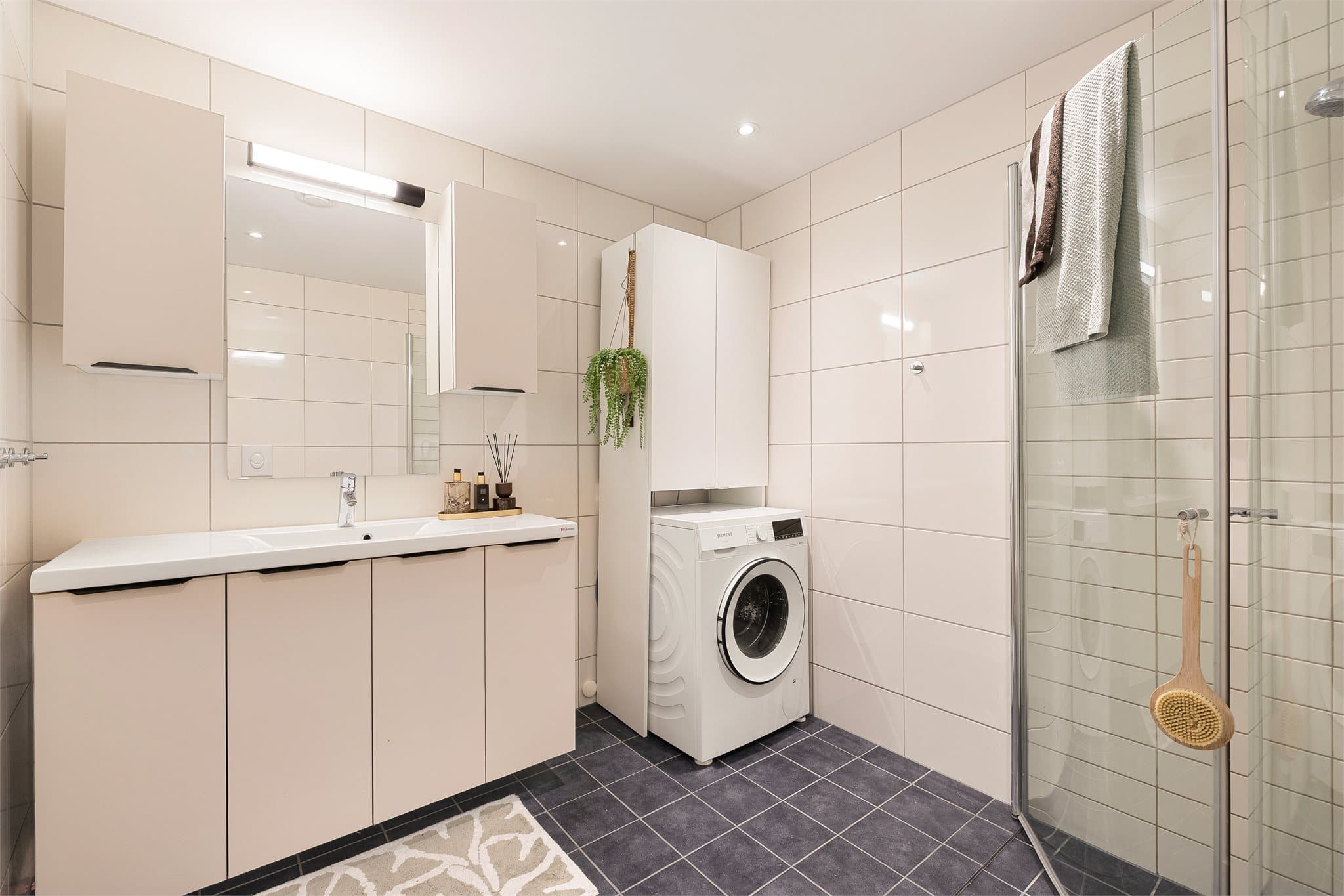 Lyst baderom med stor servantinnredning, dusjhjørne med innfellbare dører, toalett og opplegg for vaskemaskin.