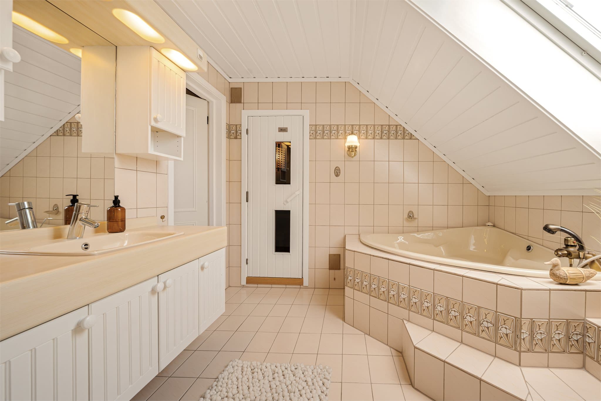 Badet i loftsetasjen kombinerer komfort og velvære med innfliset hjørnebadekar, egen badstue og praktisk dusjløsning. Lyse fliser og skråtak gir rommet et tidløst og innbydende uttrykk.