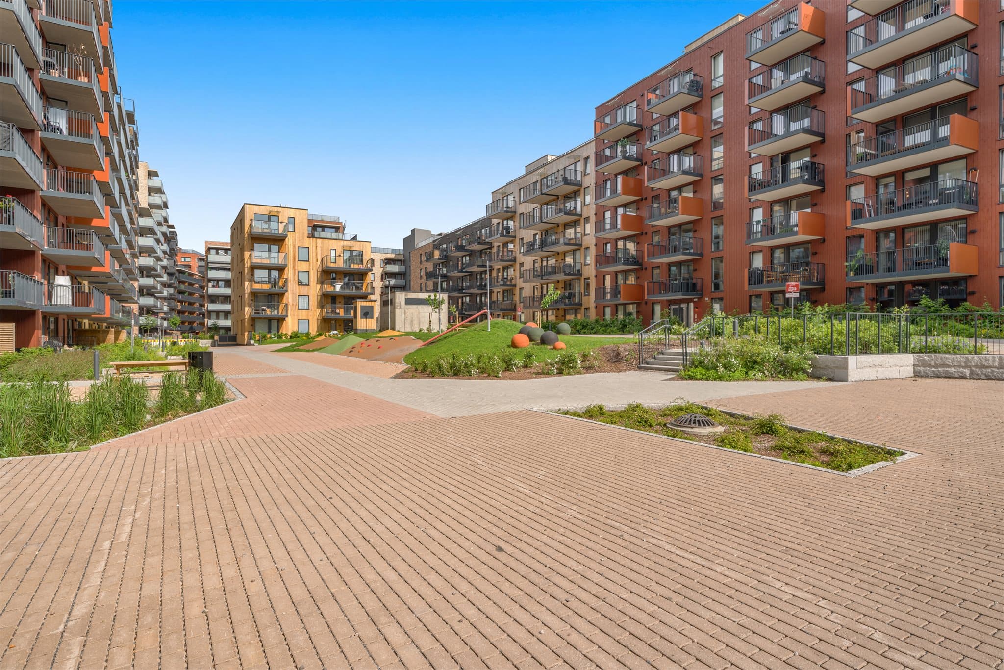 Bredt, bilfritt gårdsrom med hellelagte gangveier og moderne arkitektur.