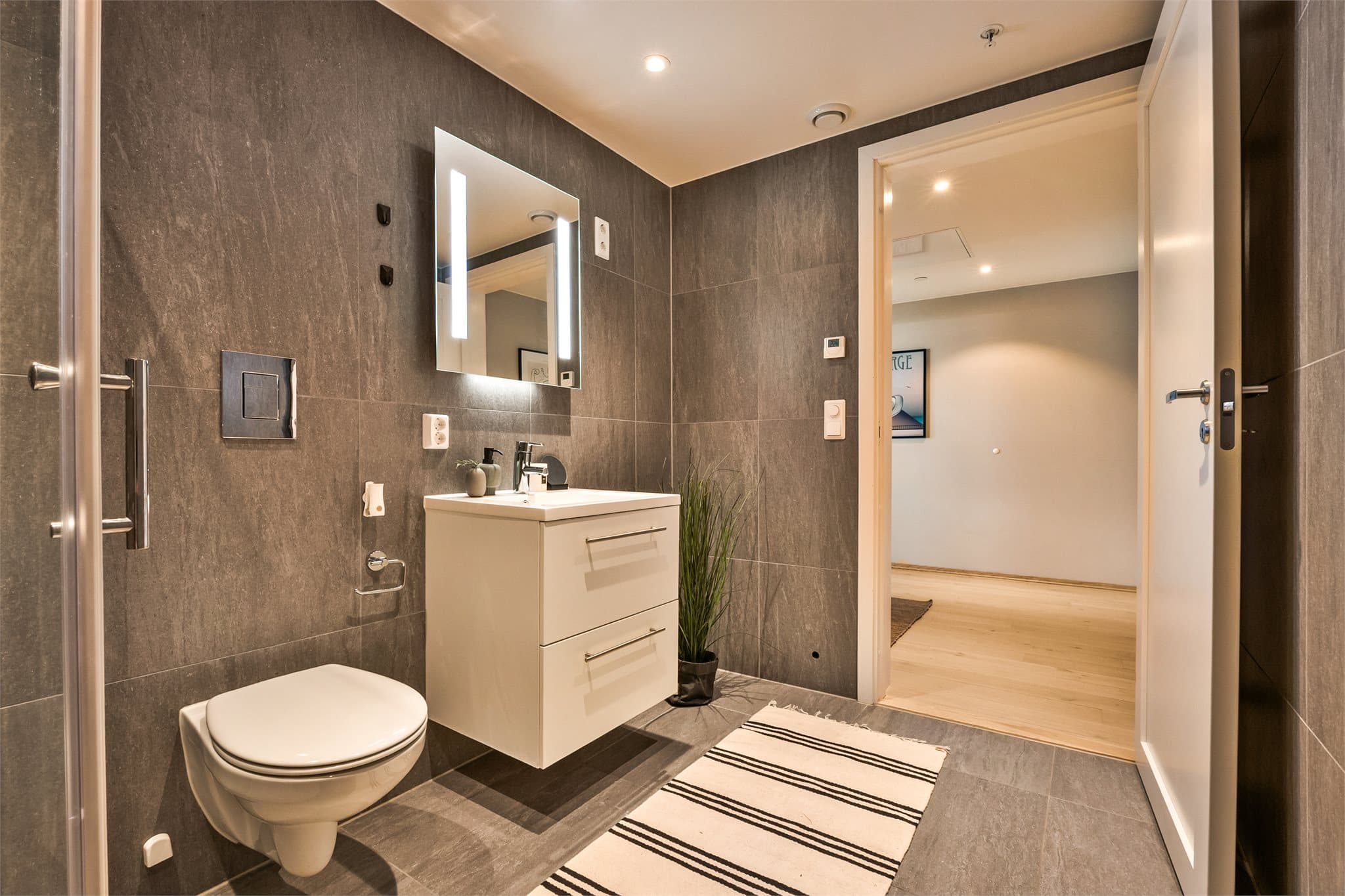 Badet er innredet med helservant på skuffeseksjon, vegghengt toalett, dusjhjørne og opplegg for vaskemaskin.