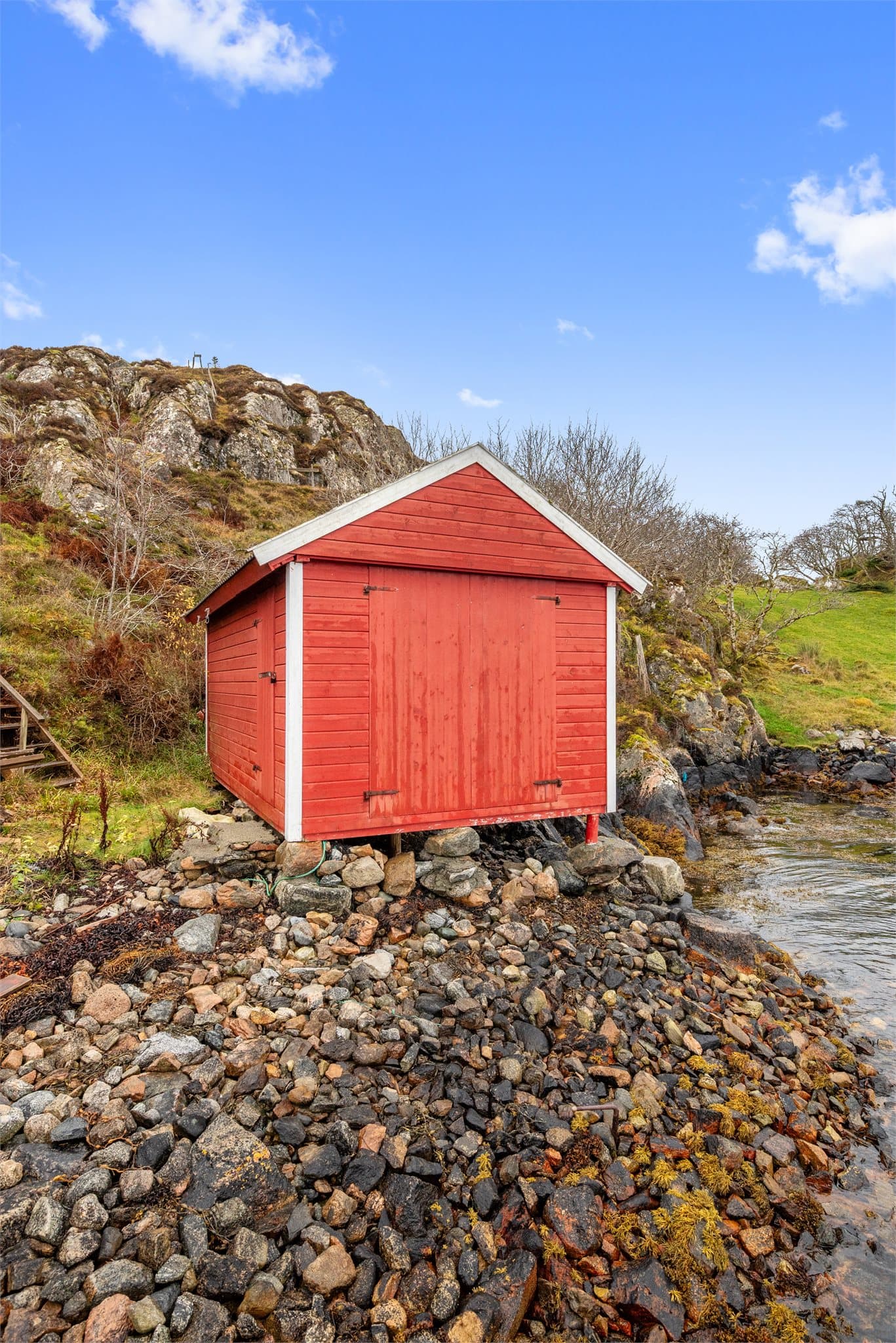 Frittstående naust ved sjøen med natursteinsfundament og stående trekledning.
