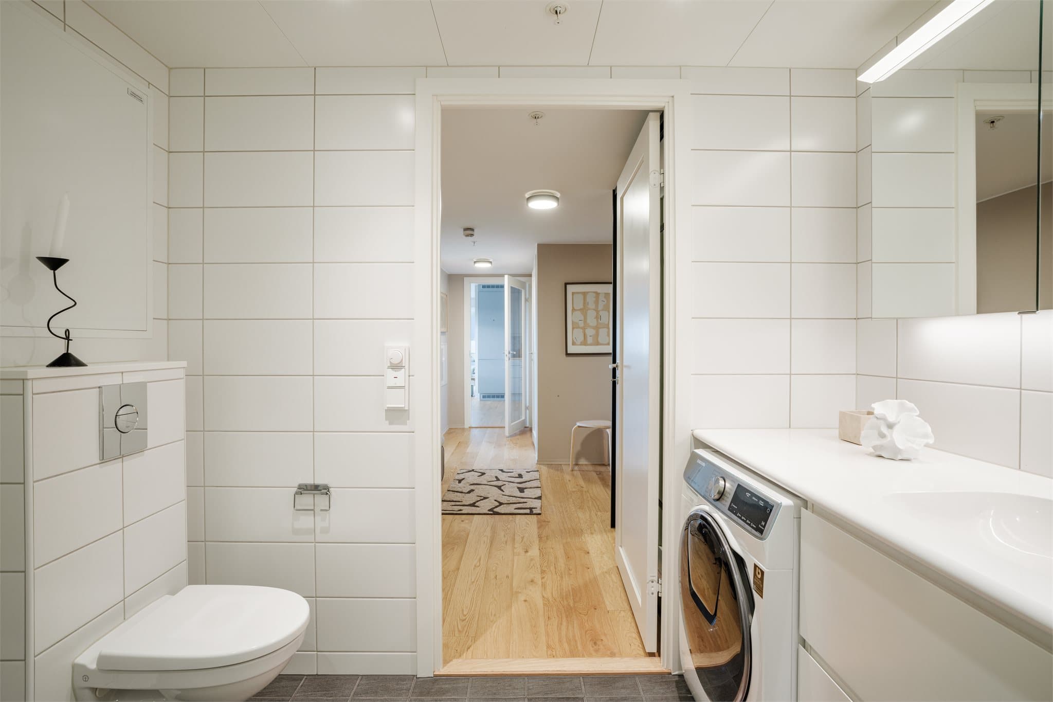 Dette badet er innredet med hvite fliser, servant med skuffer og speil med lys over.