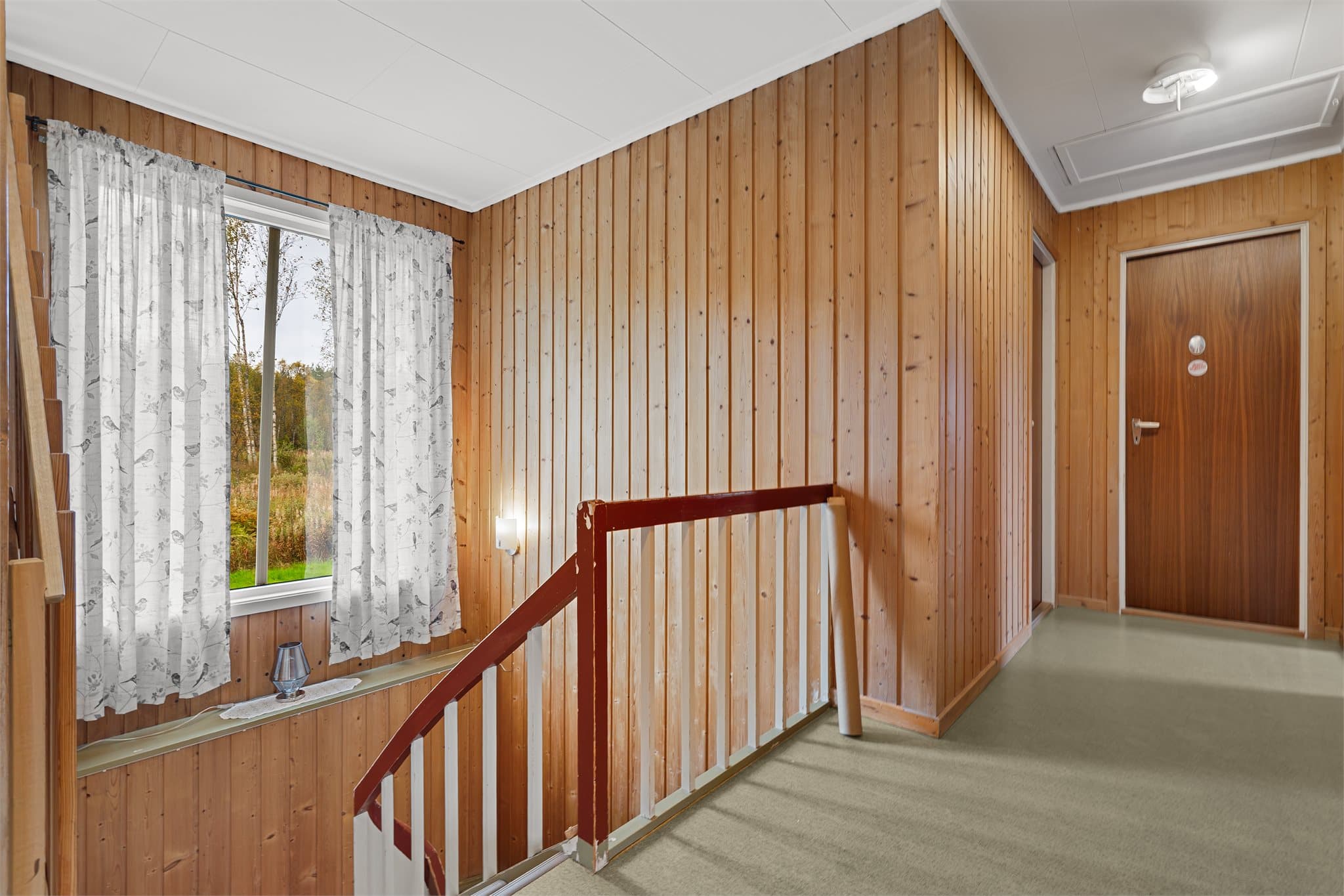 Gang i hovedetasjen kledd med trepanel og trappeløp ned til underetasjen.