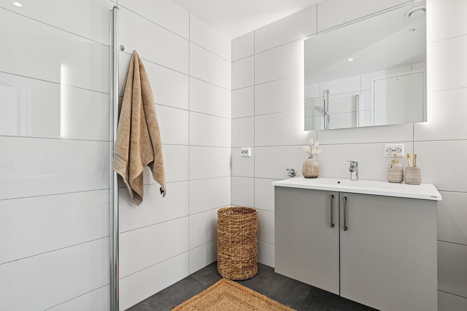 Innbydende baderom utstyrt med dusjhjørne med innfellbare dusjdører, vegghengt toalett, servantinnredning fra HTH, speil med lys og opplegg for vaskemaskin.