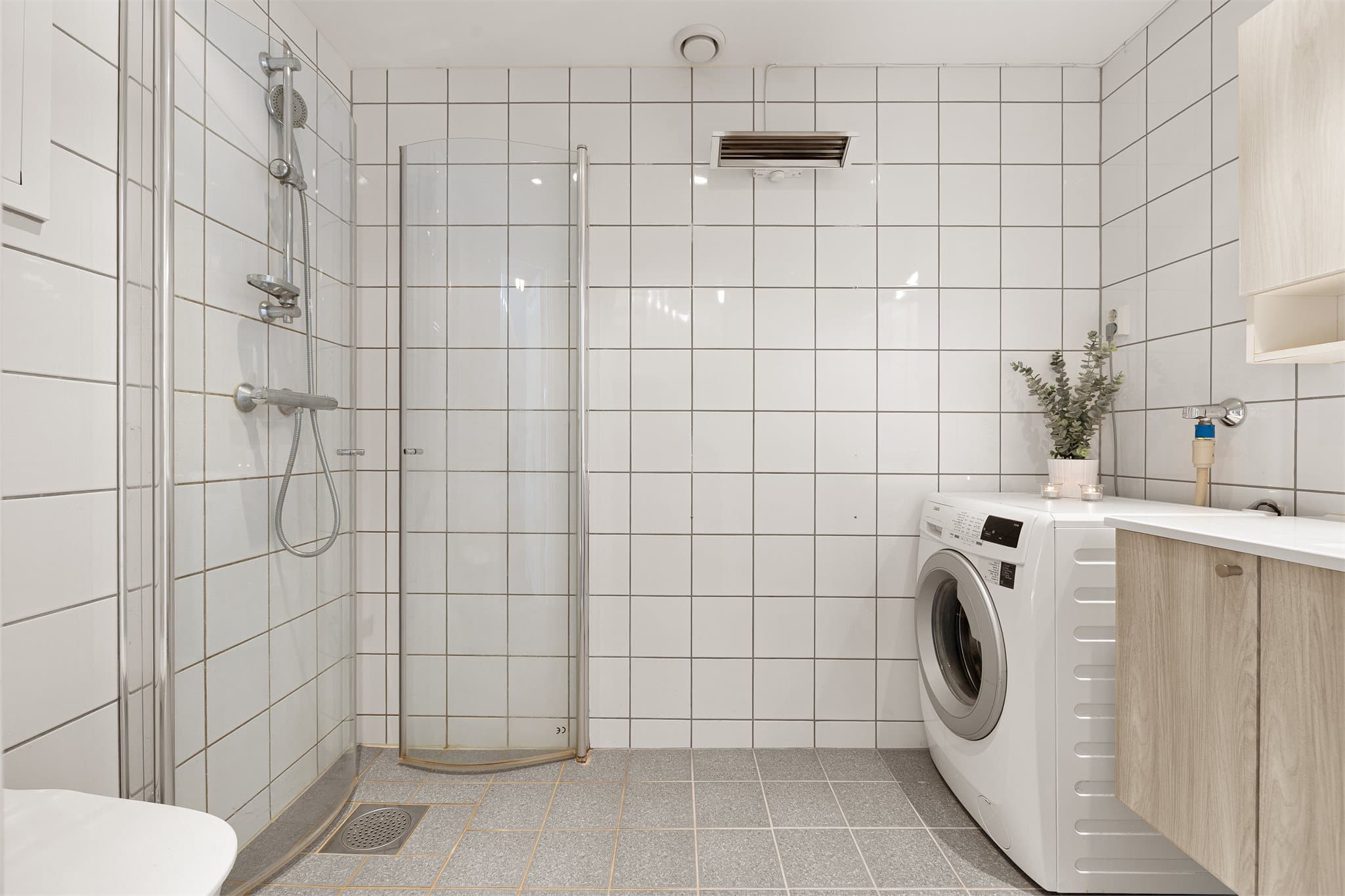 Lyst og flislagt baderom utstyrt med toalett, servantinnredning, dusjhjørne og opplegg for vaskemaskin.
