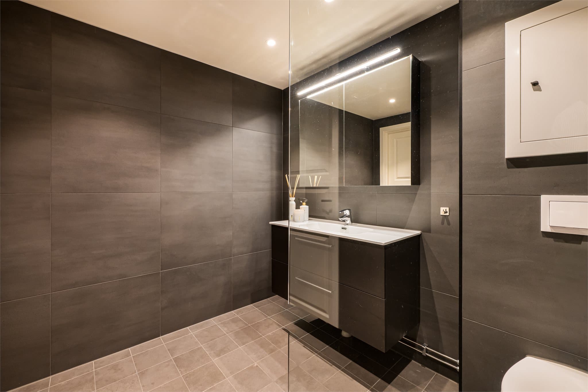 Rommet er innredet med servantskap med heldekkende servant, speilskap med integrert belysning, vegghengt toalett og dusjhjørne med glassfelt, regnfallsdusj og hånddusj.