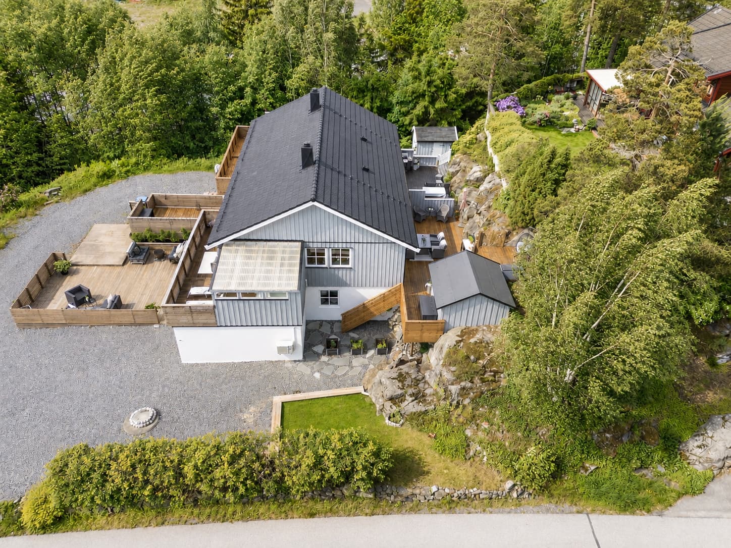 Dronefotoet viser på en flott måte hvordan boligen ligger på en romslig og lettstelt tomt med flere skjermede uteplasser, gode solforhold og naturskjønne omgivelser. Her kan du nyte både morgenkaffen og sene sommerkvelder.