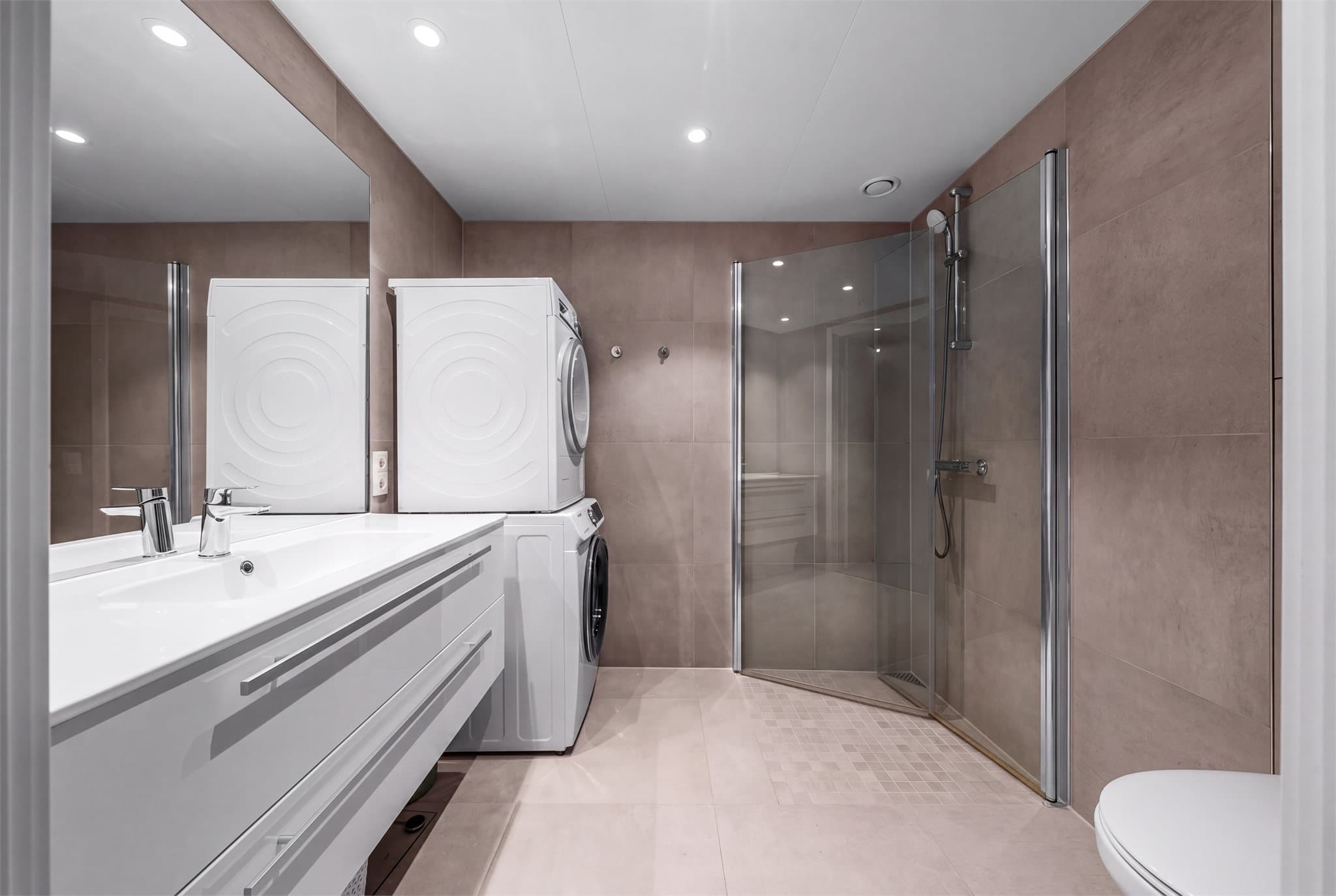 Badet er romslig og moderne utført med store fliser i gråtoner på gulv og vegger, gulvvarme og downlights i tak.