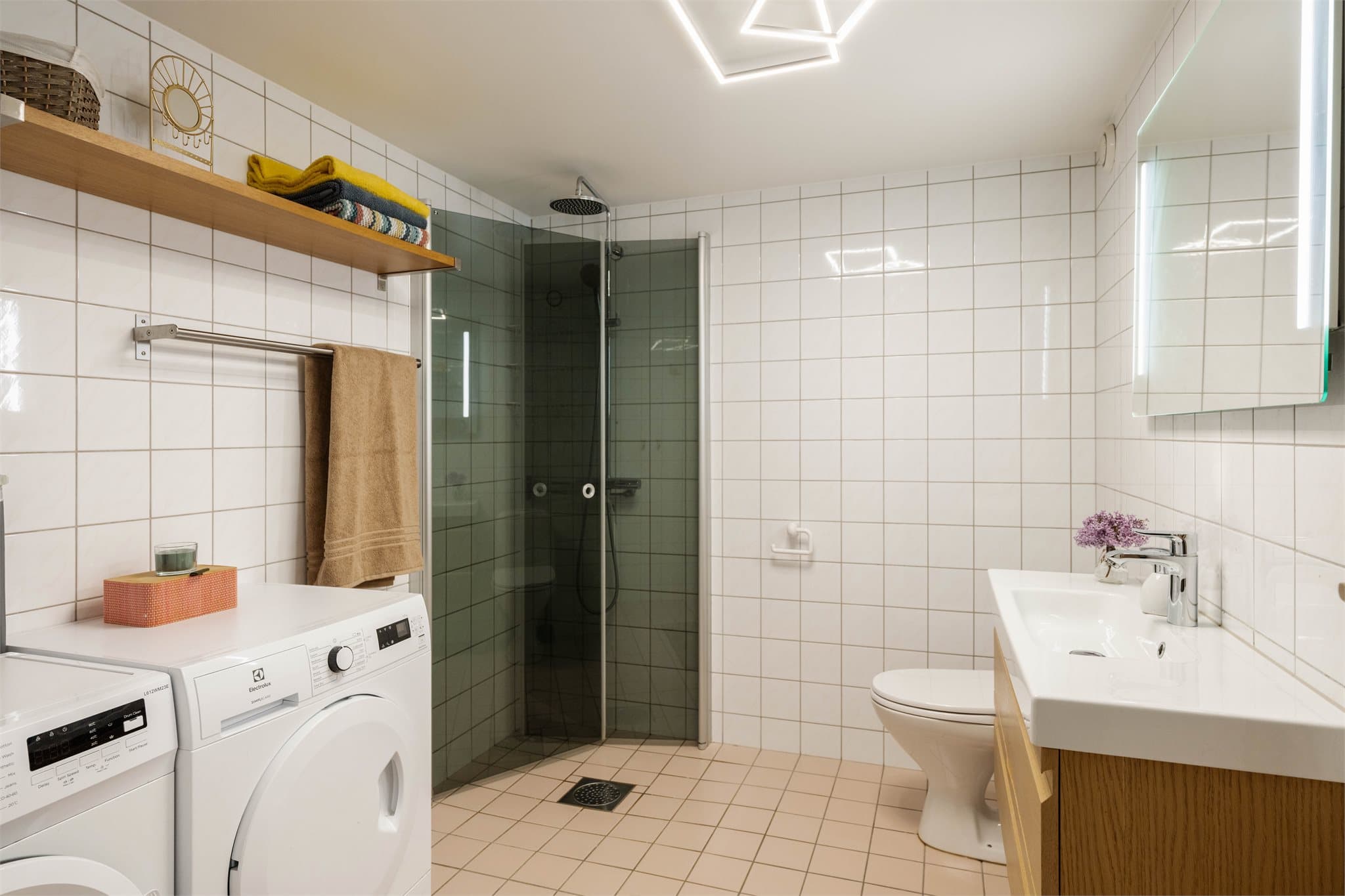Romslig baderom med dusjhjørne med svingdør, servantskap i glatt utførelse med heldekkende servant, speil med integrert LED-lys og WC.