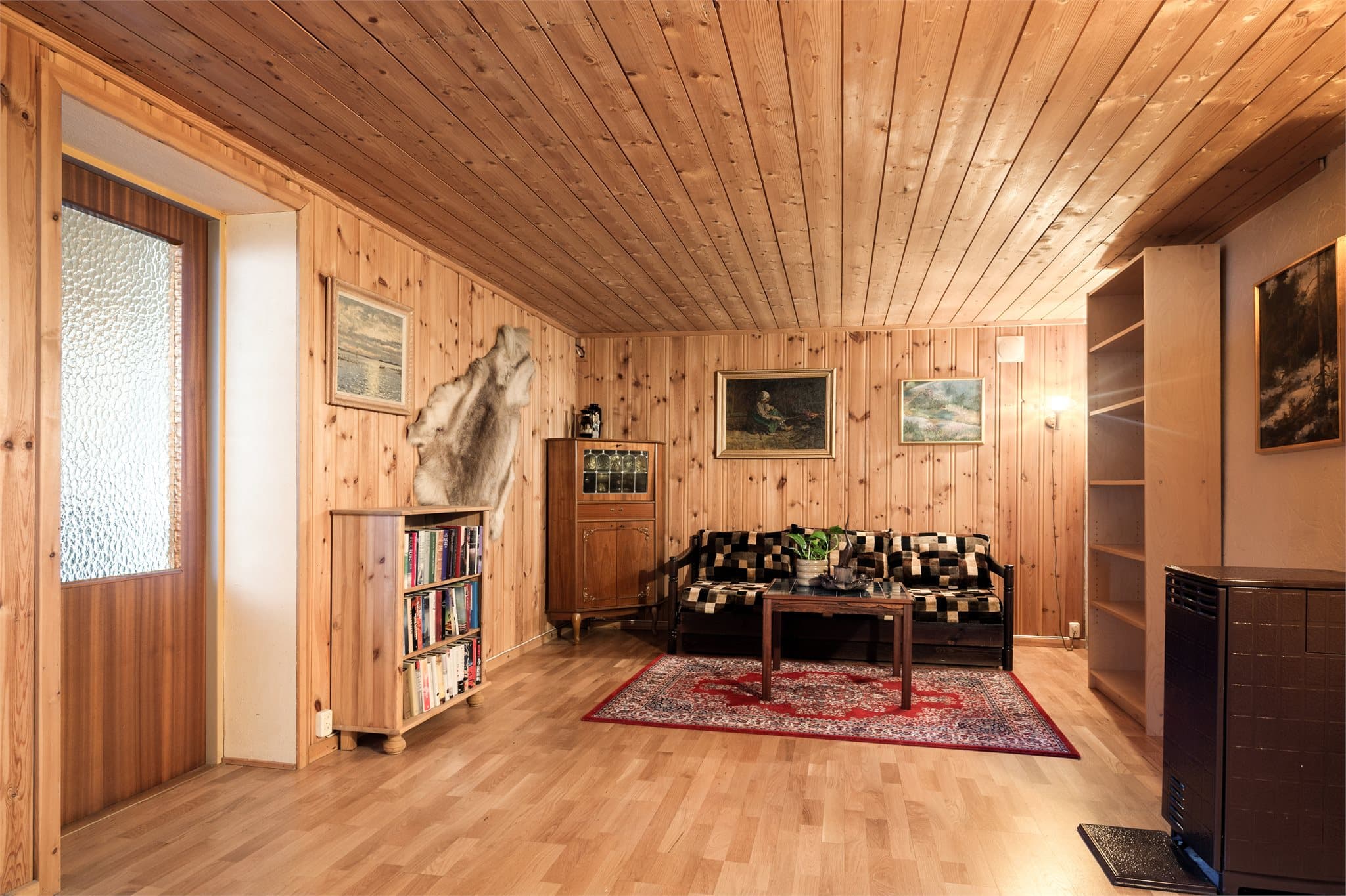 Veggene er kledd i klassisk furupanel, og taket følger opp med panel som gir rommet en lun og helhetlig ramme. Laminatgulvet gjør det lettstelt og anvendelig, mens vedovnen sørger for ekte varme og stemning når kulden setter inn.