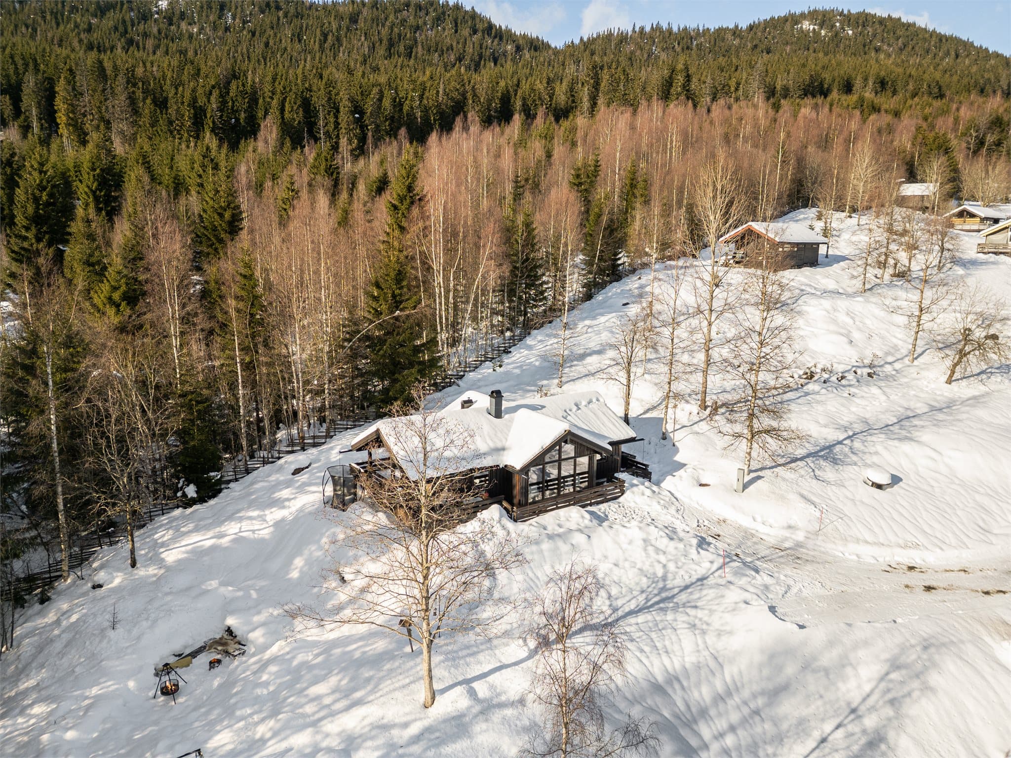 Fra luften fremheves den harmoniske plasseringen i terrenget, med åsrygger og skogsområder som omkranser hytta og skaper en følelse av privatliv og ro. Hytta skiller seg tydelig ut med sin moderne arkitektur, store glassflater og brunbeisede fasade.