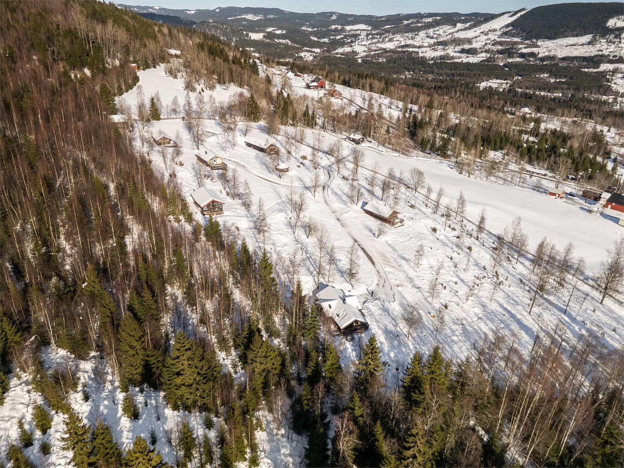 Droneperspektivet viser også den naturskjønne utsikten mot Hurdal Skisenter og Hurdalsjøen i det fjerne, samt den overbygde verandaen, bålplassen og uteområdene som skaper perfekte rammer for hytteliv året rundt.