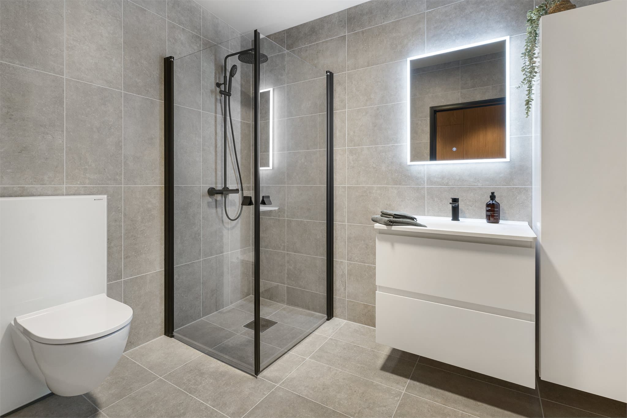 Bad i hovedetasjen: Badet er utført med flislagte vegger og gulv med vannbåren varme. Innredningen består av veggmontert Geberit-toalett og dusj med hengslede glassdører. Toalettet er forberedt for eventuell senere montering av dusjtoalett.