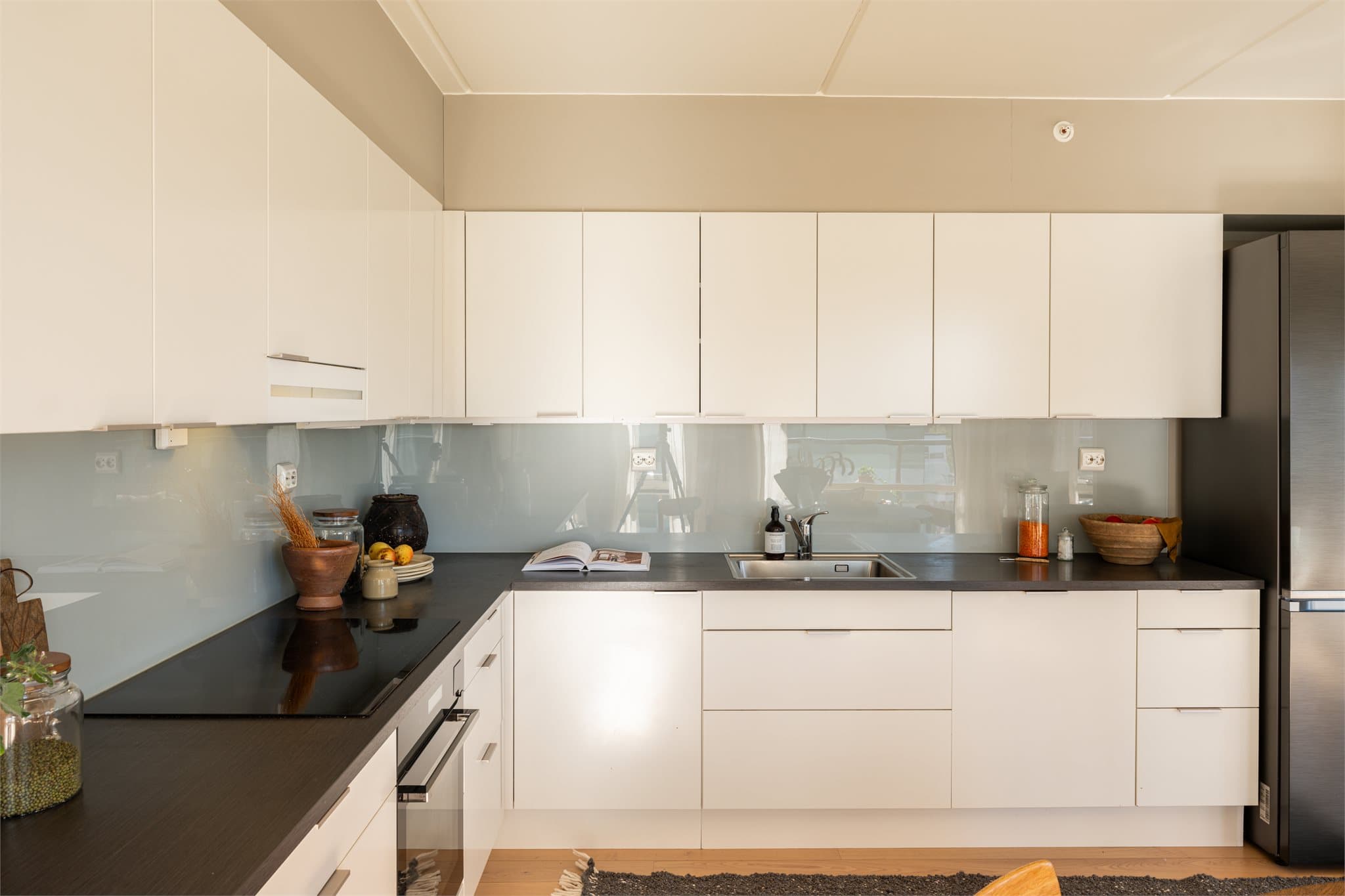 Kjøkkenet er utformet i L-form med hvit, glatt innredning, mørk laminatbenkeplate med stålvask og elegante, spesialtilpassede glassfelt over hele benken.