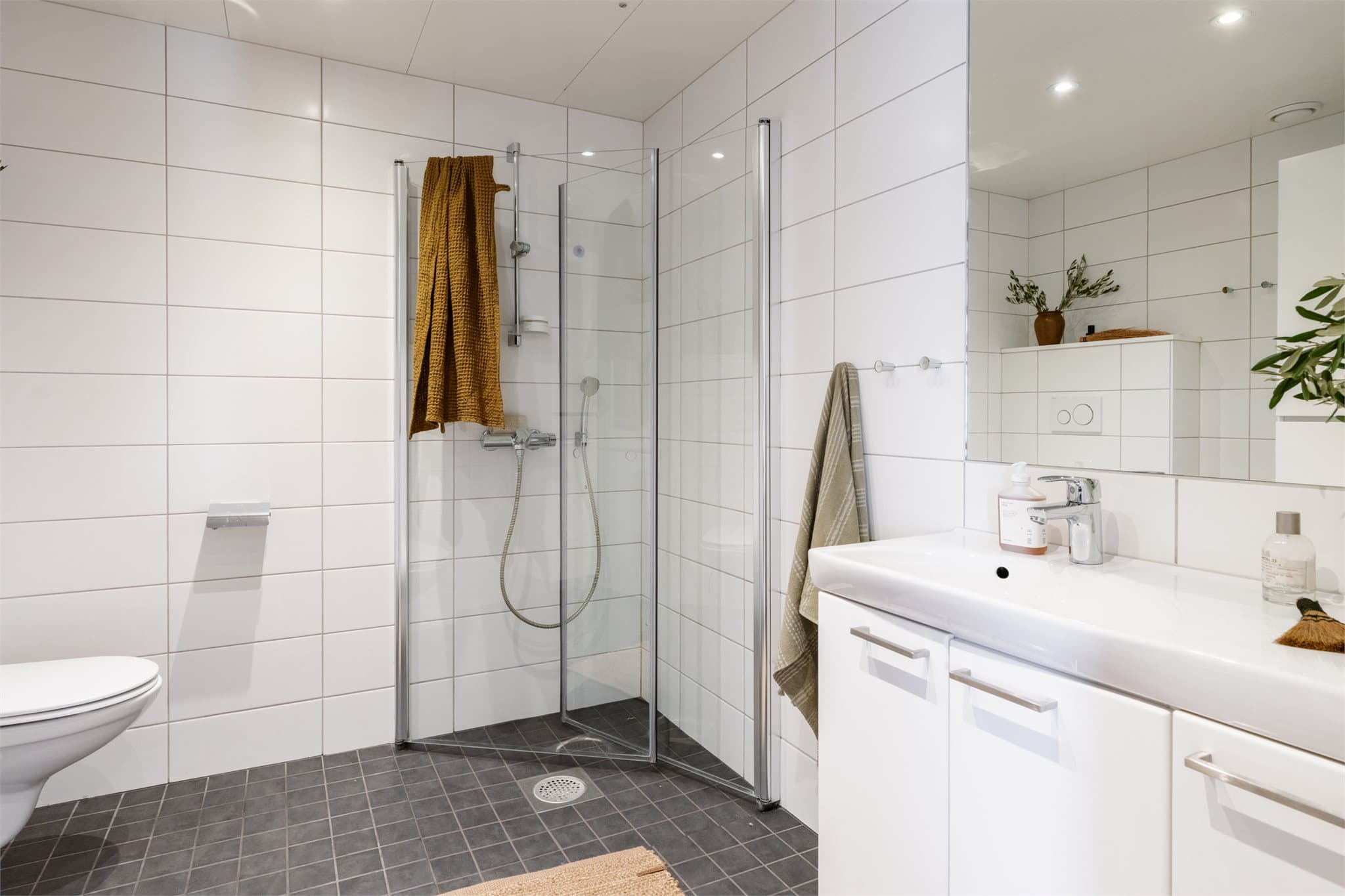 Innredningen består av servant med skap, speil, høyskap, vegghengt toalett og dusjhjørne med glassdører.