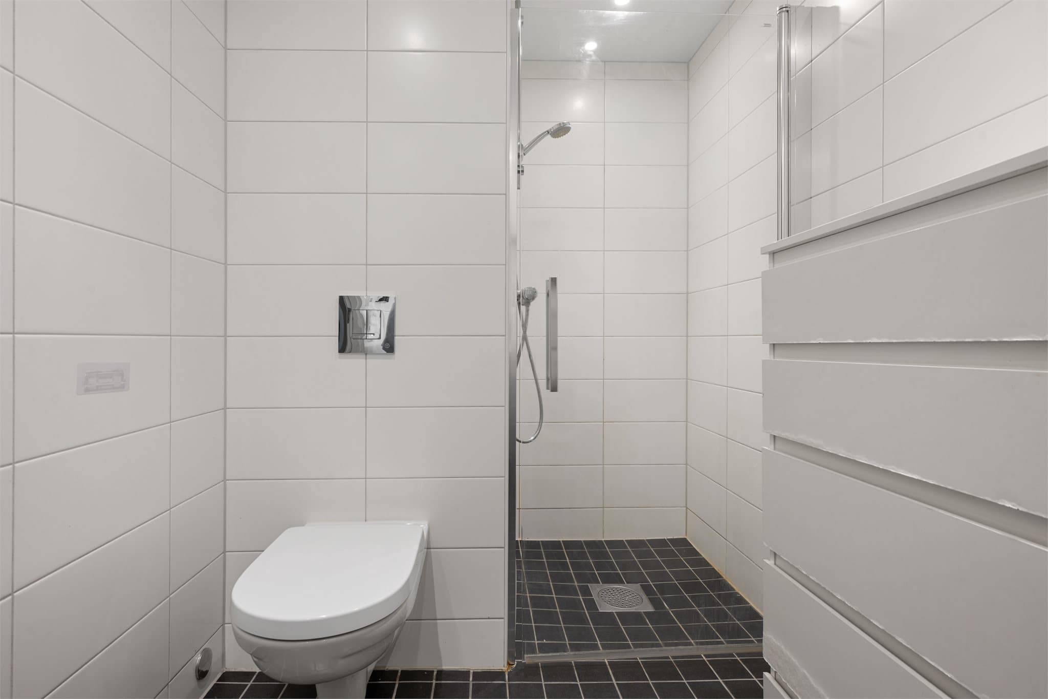 Flislagt bad med vegghengt toalett, dusjløsning og servant på skuffeseksjon.