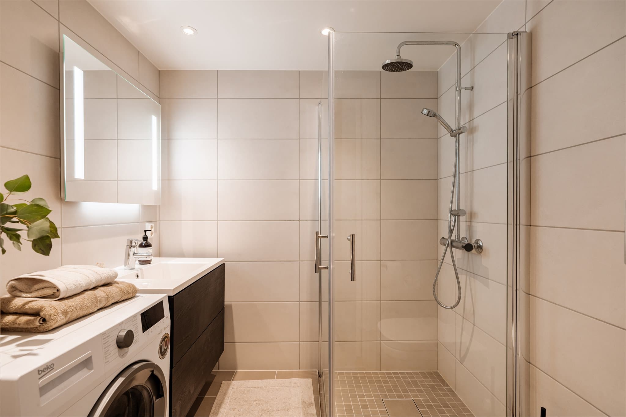 Moderne, flislagt bad med en slett innredning med heldekkende servant, speil med integrert lys opplegg for vaskemaskin.