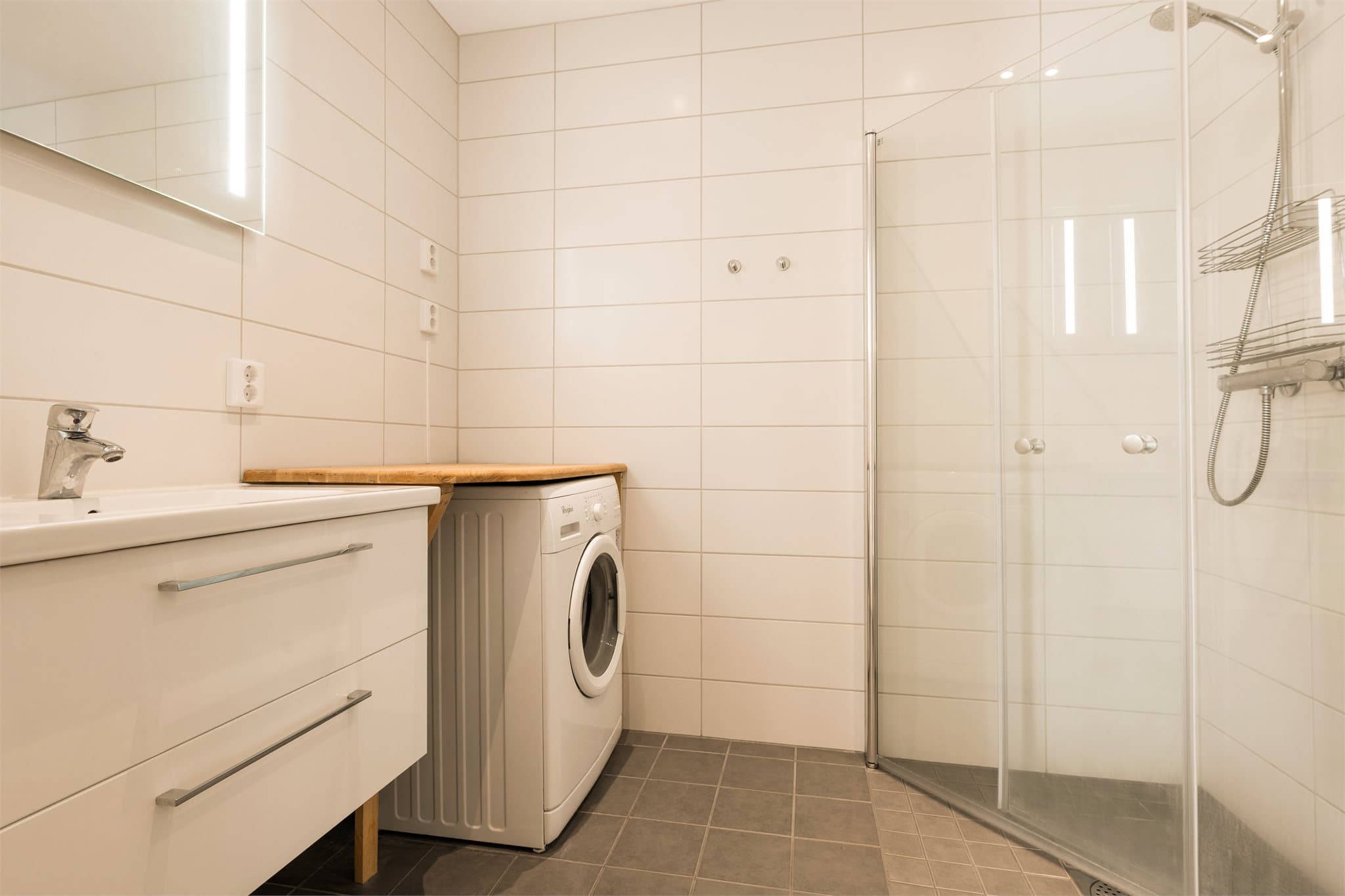 Badet er innredet med dusj på vegg og dusjdører i herdet glass, helservant på skuffeseksjon, speil med belysning og klosett.