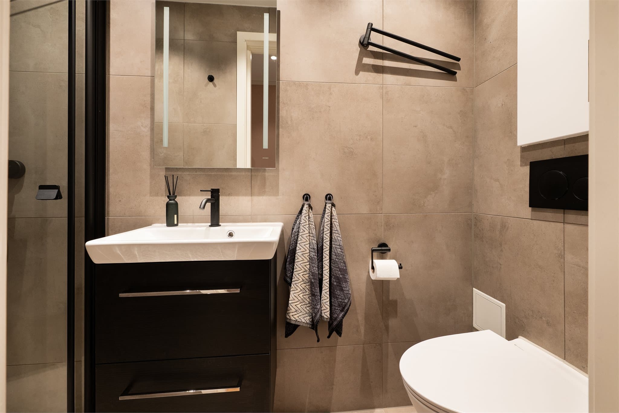 Rommet er innredet med servantskap med glatte, fabrikklakkerte fronter og heldekkende servant. Det er speil med belysning på vegg over servant. Videre er det veggmontert toalett og dusjhjørne med foldedør, regnfallsdusj og håndholdt dusj.