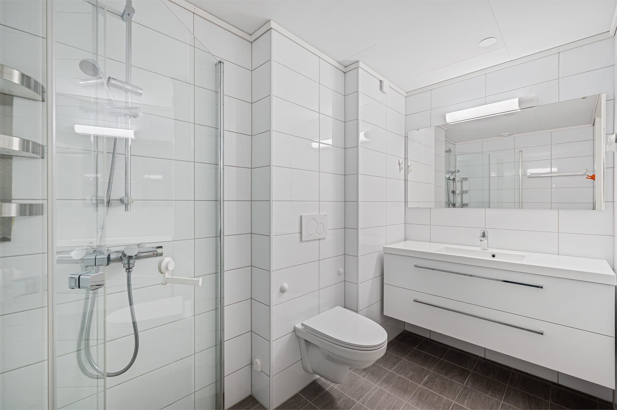 - Badet er innredet med heldekkende servant, speil, vegglampe, vegghengt toalett og dusj med innfellbare dører i herdet glass samt dusjgarnityr med hånddusj -