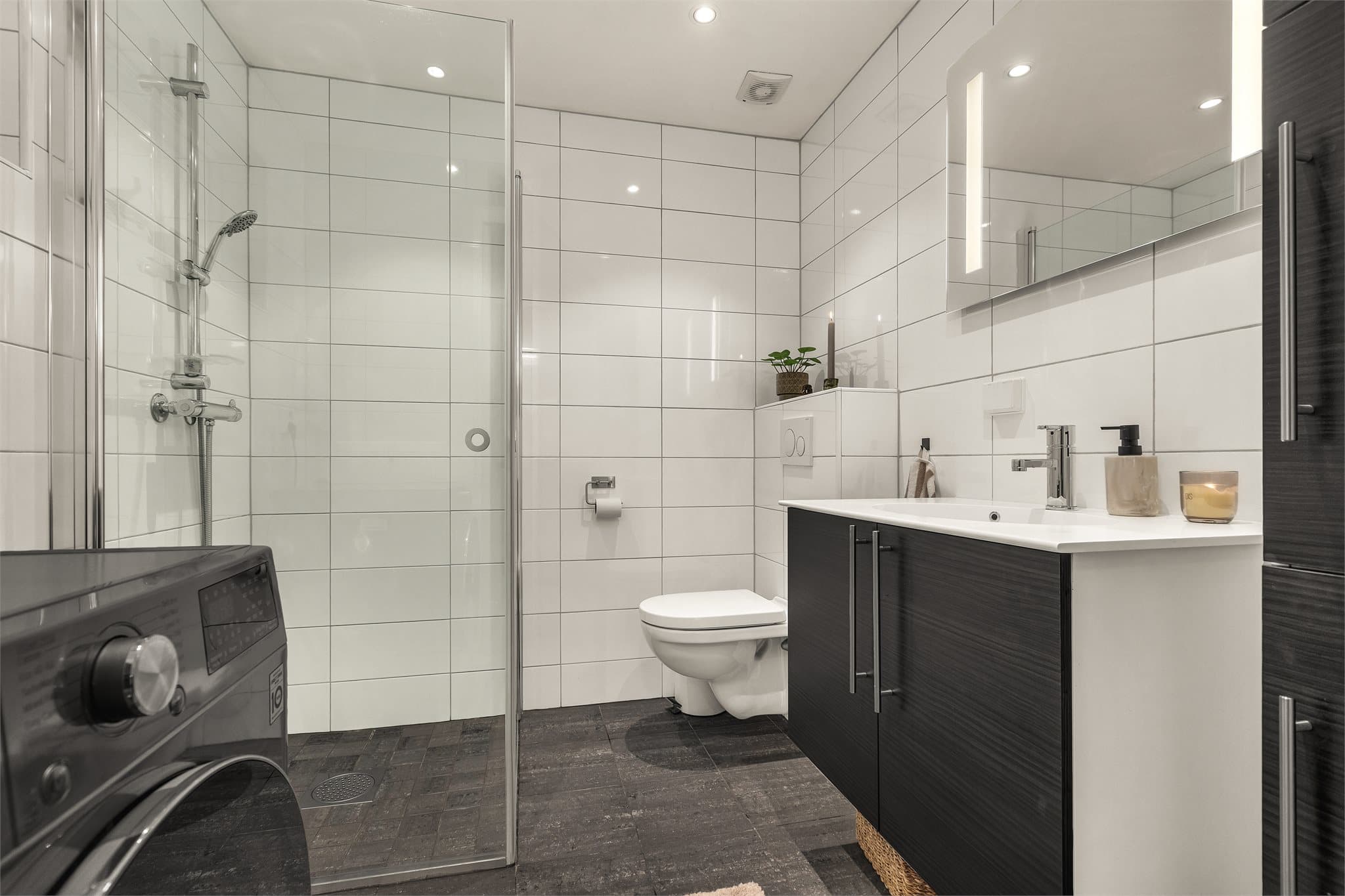 Badet har flislagte vegger, mørke gulvfliser med varmekabler og dusjhjørne med glassvegg.