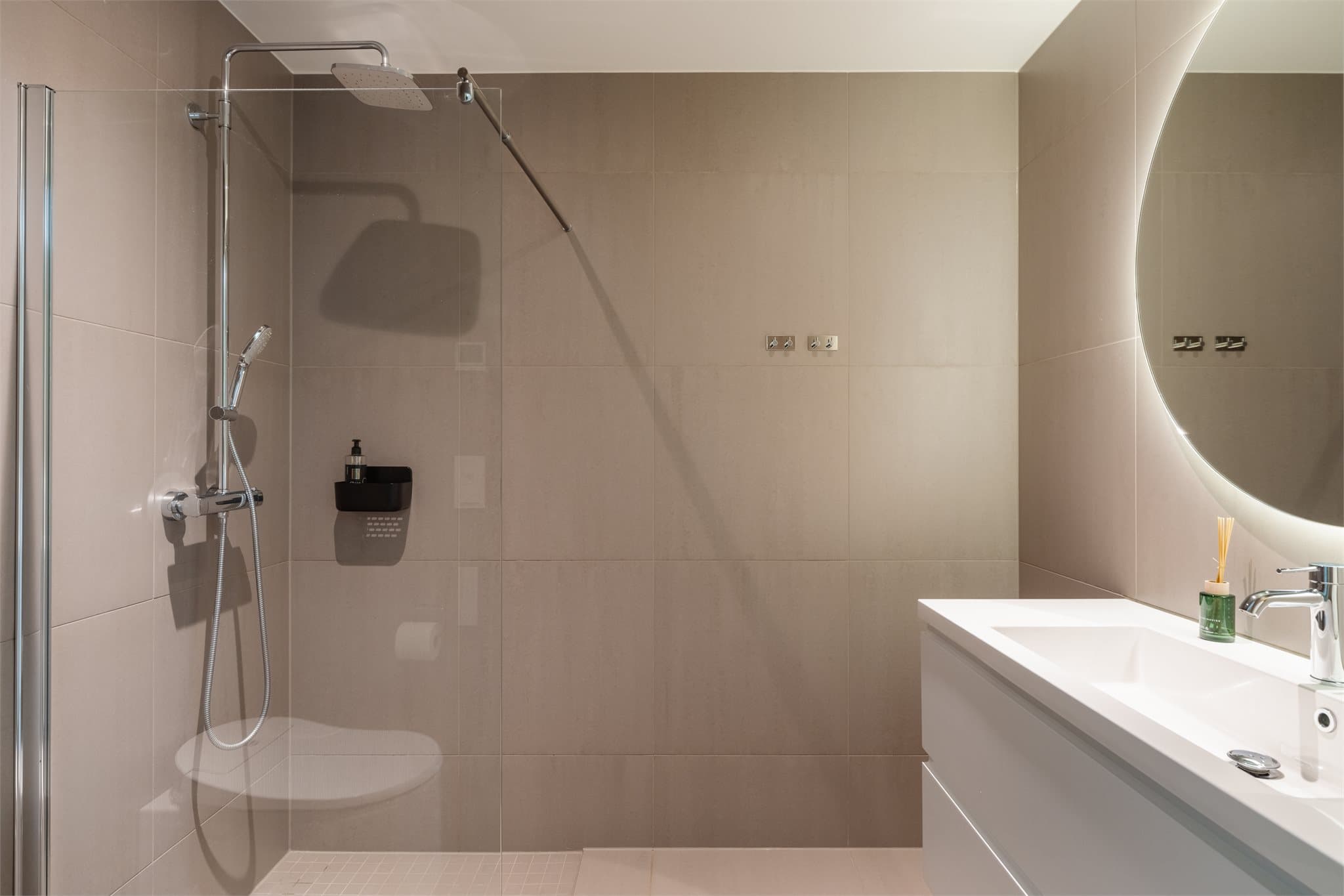 Badet tilknyttet hovedsoverommet har et moderne og gjennomført uttrykk med store fliser på vegger og gulv, supplert med mindre mosaikkfliser i dusjsonen. Den behagelige ton-i-ton-paletten gir et harmonisk preg.