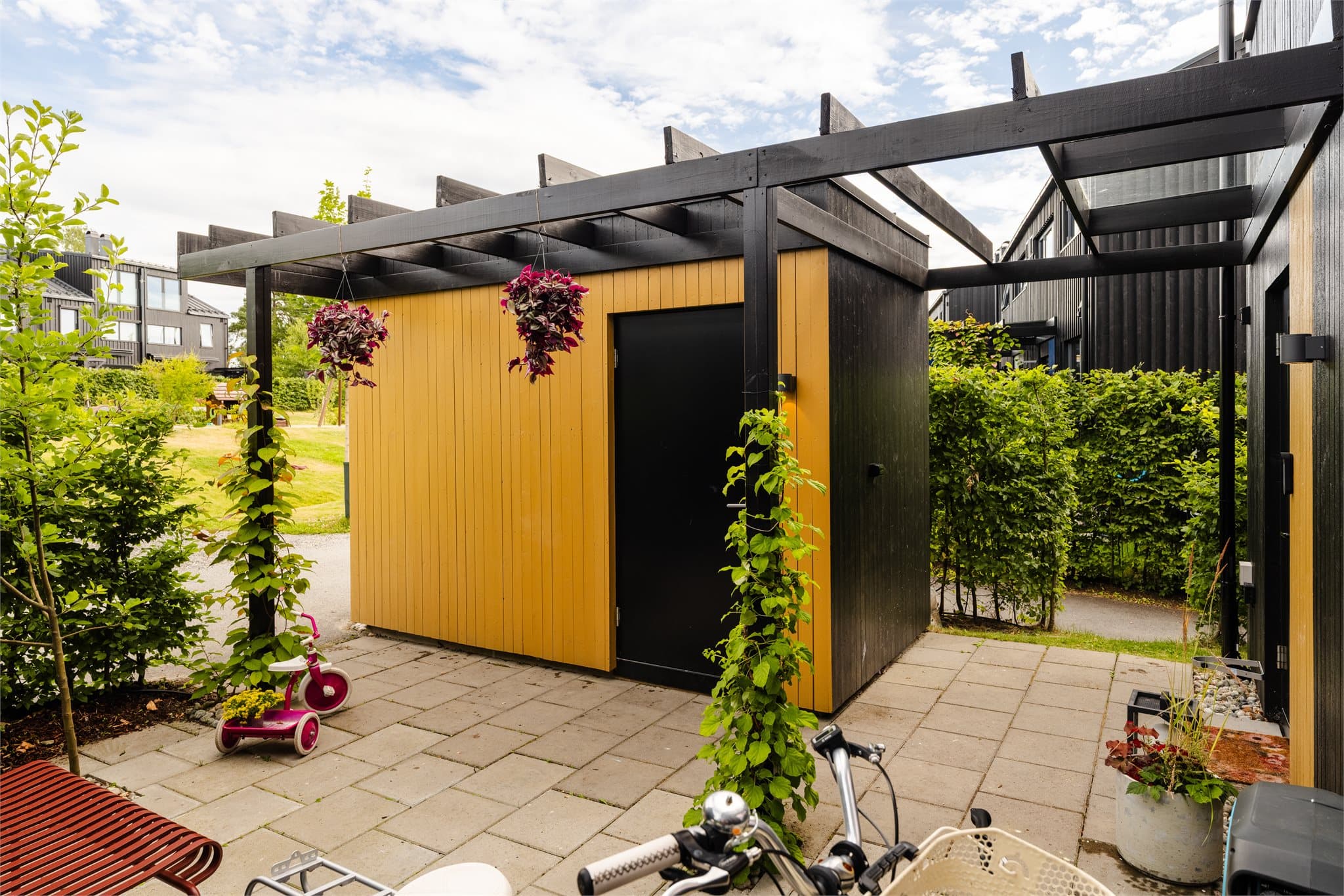 Den fargede inngangsdøren står i fin kontrast til den mørke fasaden, mens pergola og frodig beplantning rammer inn inngangssonen.