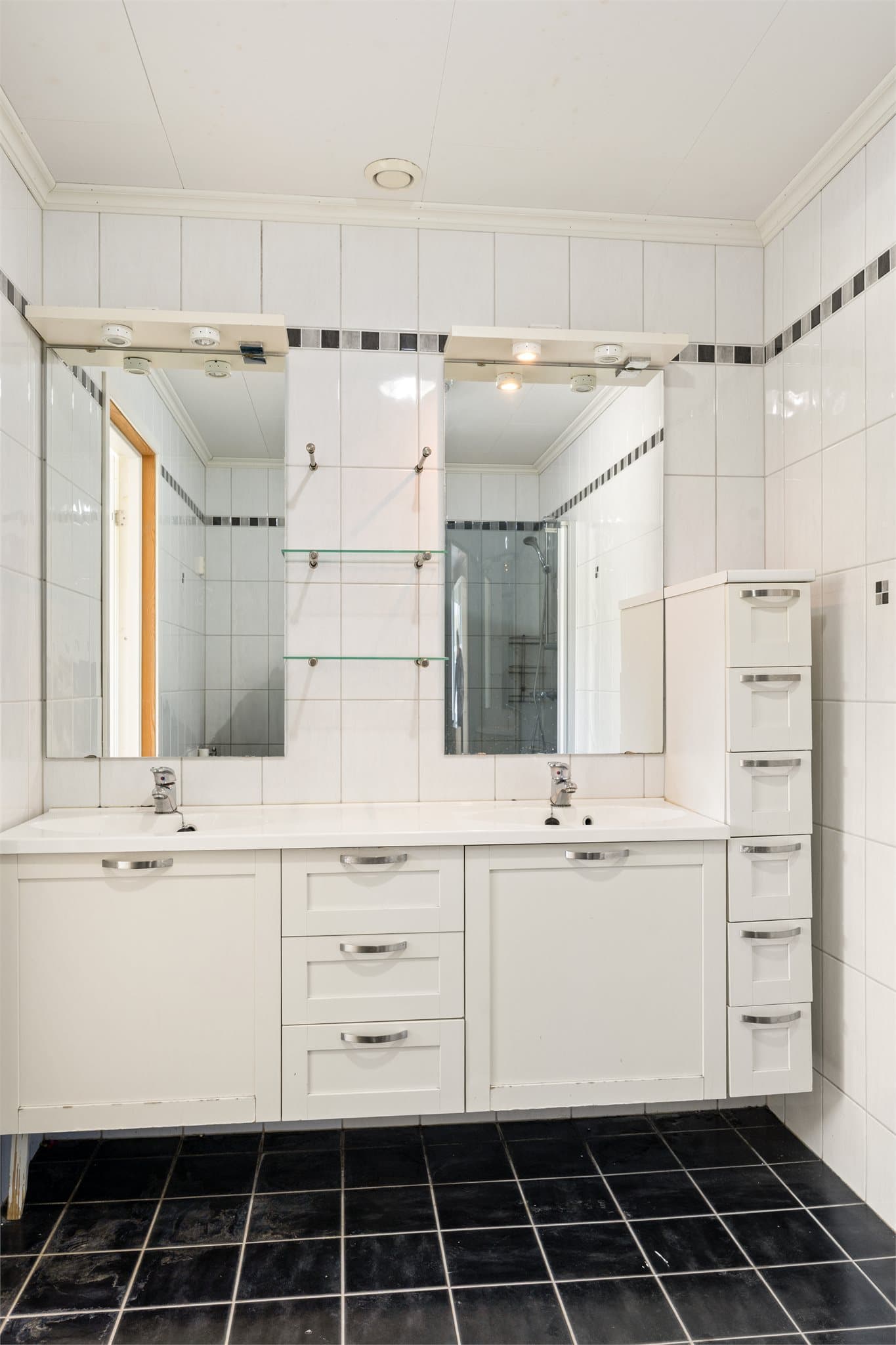 Videre finner du stående klosett samt en praktisk dobbelservant med tilhørende doble speil og belysning, integrert i en hvit, profilert baderomsinnredning.