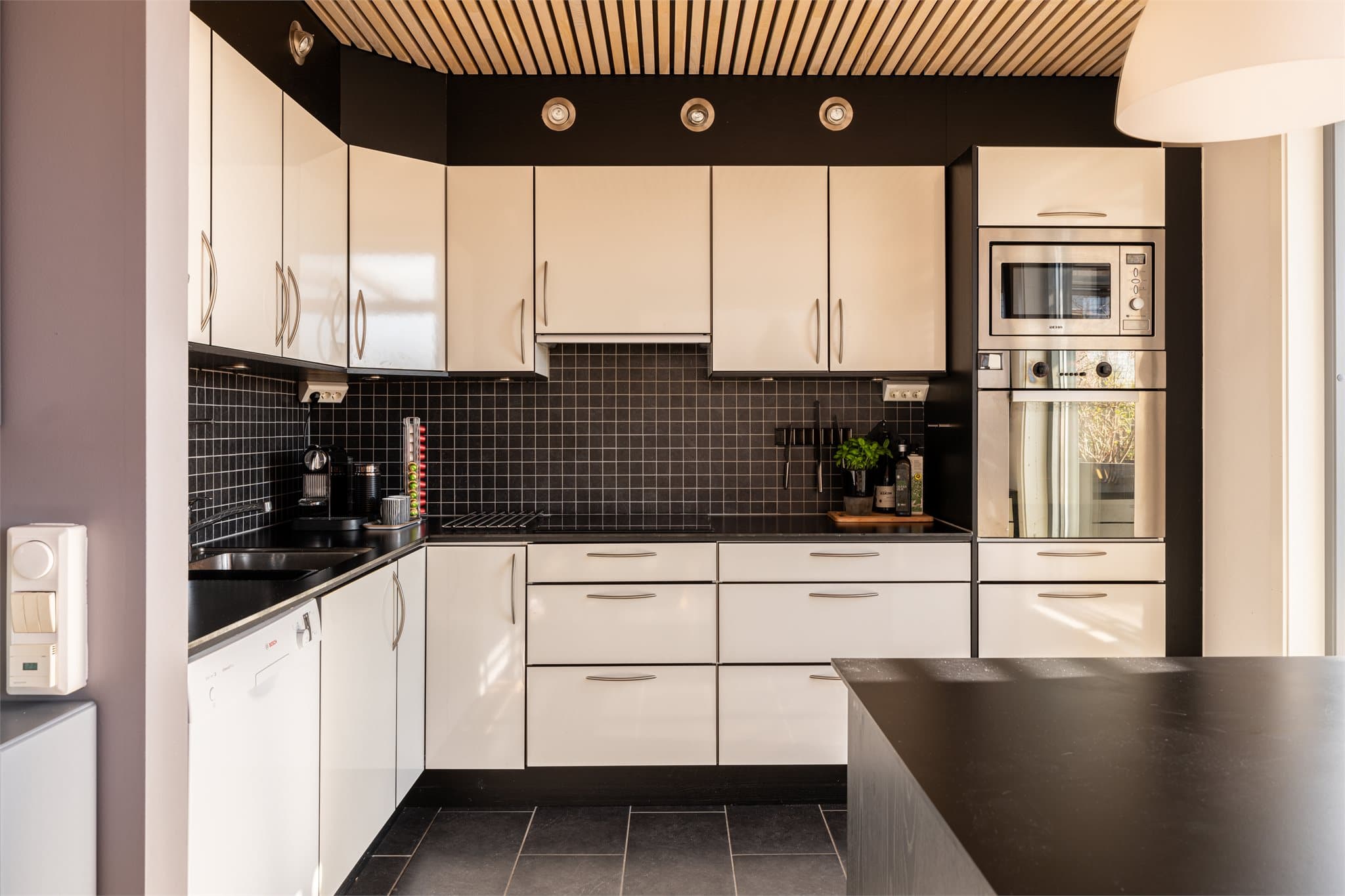 Kjøkkeninnredning med glatte, lyse fronter og mørk benkeplate og fliser.