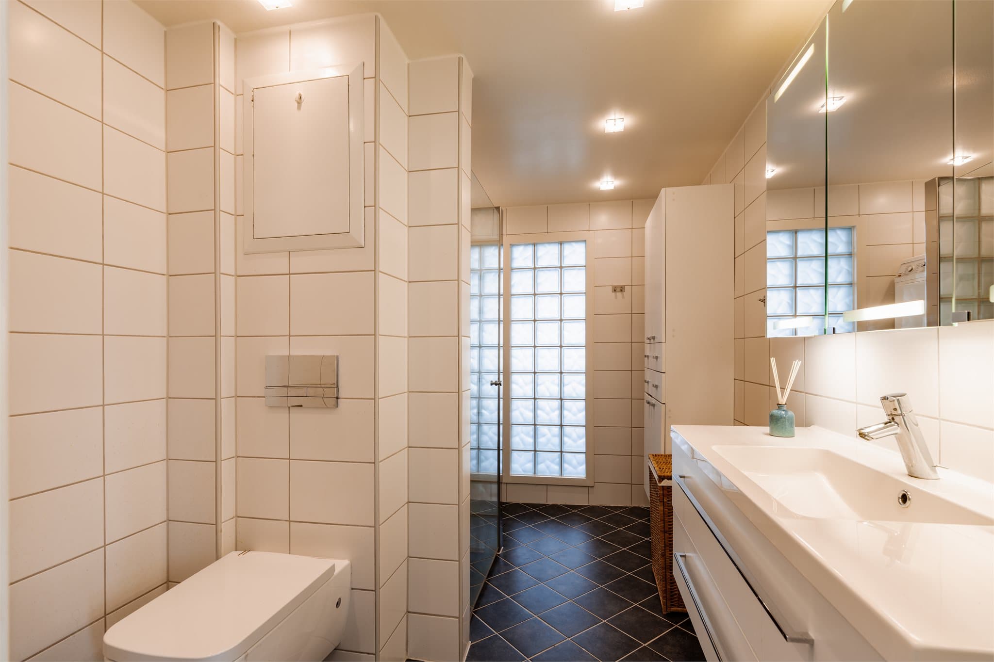 Flislagt bad med varmekabler i gulv. Innredningen består av benkeskap og veggskap med glatte fronter, servant og vegghengt toalett med innebygget sisterne.