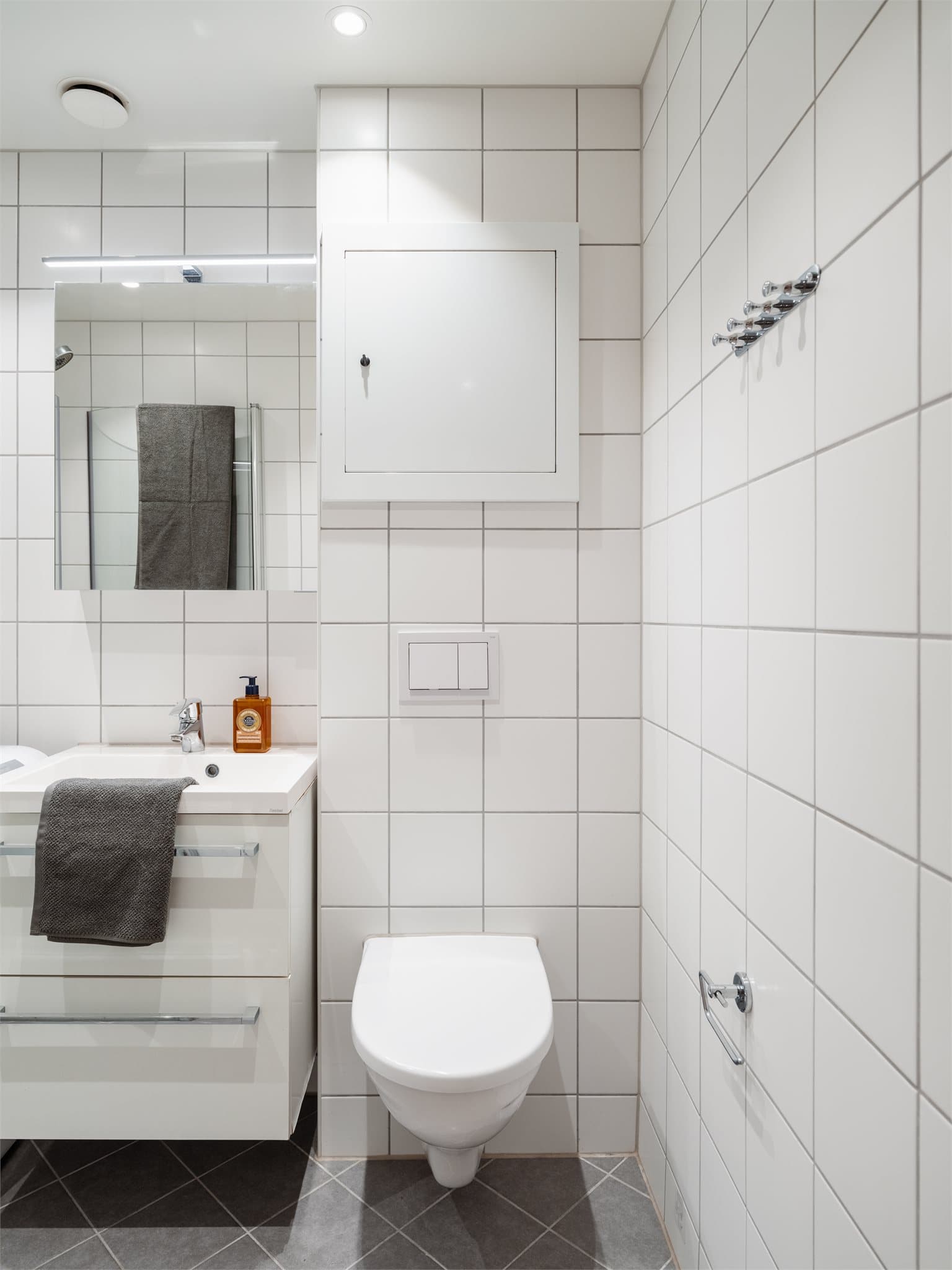 Badet er innredet med servant med skuffeinnredning, vegghengt klosett med innebygget sisterne og dusj på gulv med dusjdører i herdet glass.