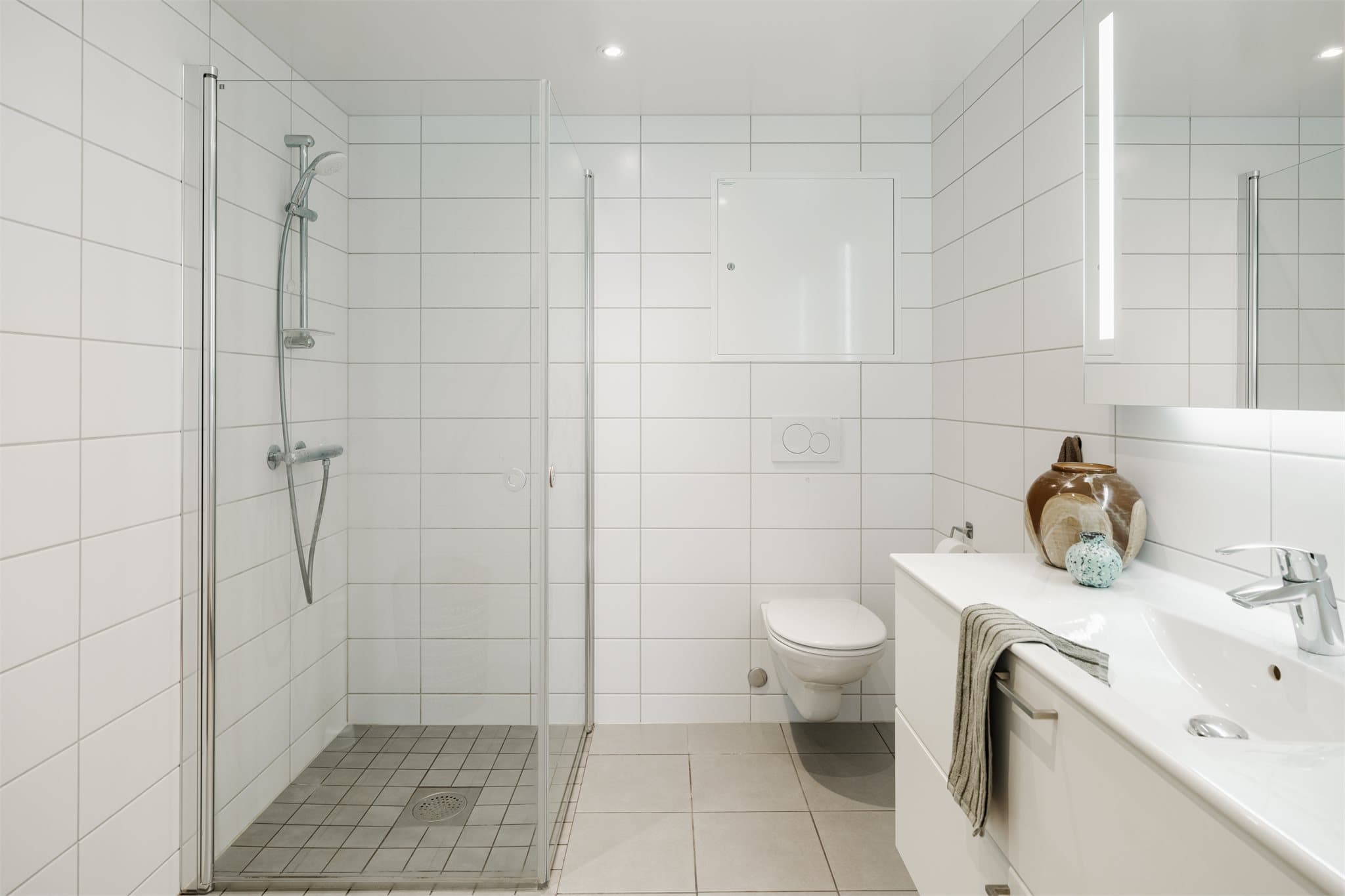 Badet er utstyrt med dusj på gulv med dusjdører i herdet glass, servant med skapinnredning samt vegghengt toalett med innebygget sisterne.