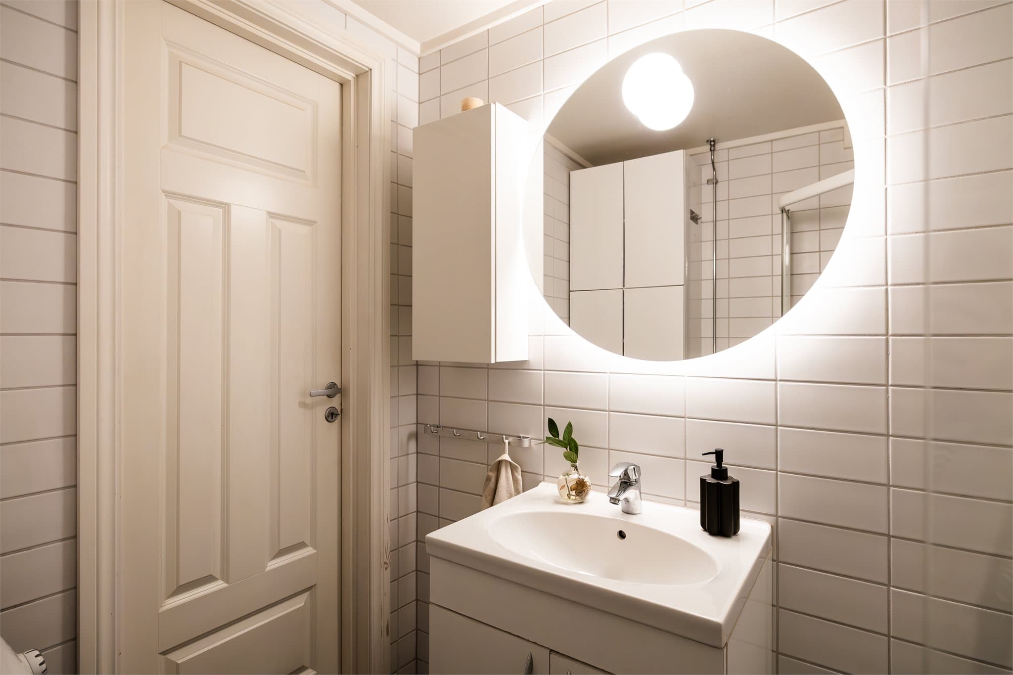 Et lyst og flislagt baderom med rundt speil og god plass til oppbevaring av toalettsaker i både servantinnredning og vegghengt skap.