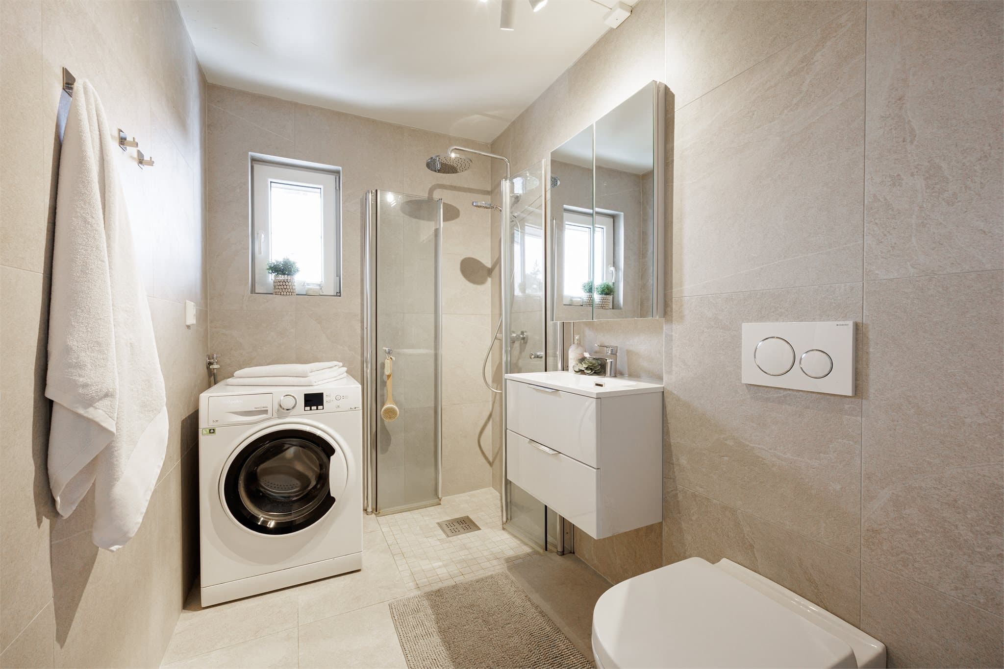 Flislagt bad med moderne innredning bestående av benkeskap med glatte fronter og veggskap med speilfronter. Vegghengt toalett med innebygget sisterne, dusj med innfellbare glassdører samt opplegg for vaskemaskin.