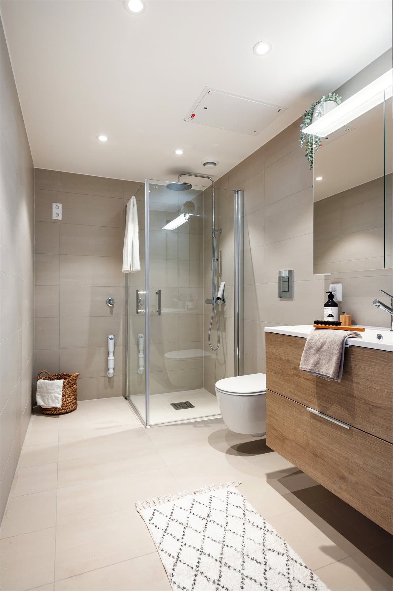 Et stilrent baderom med lune fliser og moderne innredning, hvor den nedsenkede dusjsonen er flislagt med matchende 5x5 cm fliser for et helhetlig preg, mens dusjdører med kromdetaljer setter prikken over i-en med sitt elegante uttrykk.