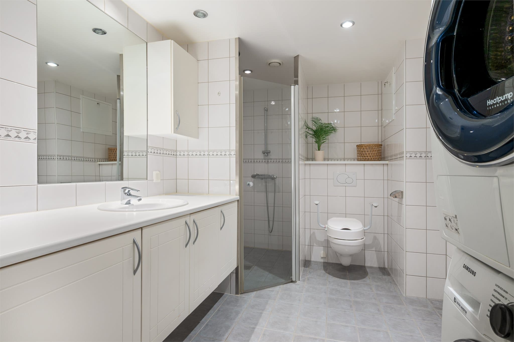 Flislagt baderom med tidløse fliser, dusjnisje, vegghengt wc og nedfelt servant på baderomsinnredning. Det er plass til vaskemaskin og tørketrommel i vaskesøyle.