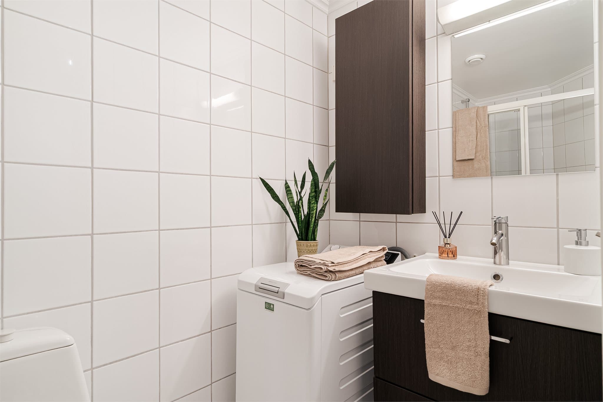 Lyst baderom med flislagte overflater, en moderne mørkebrun servantinnredning, dusjhjørne, toalett og opplegg for vaskemaskin.